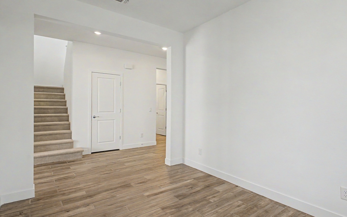 A room with light wood flooring, white walls, and a carpeted staircase leading up. Two white doors are visible in the hallway.