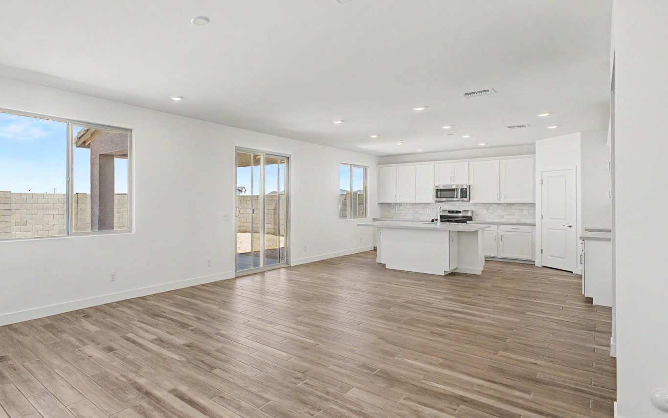 Empty modern open-plan kitchen and living area with wood flooring, white walls and cabinets, large windows, sliding glass door, and recessed ceiling lights.