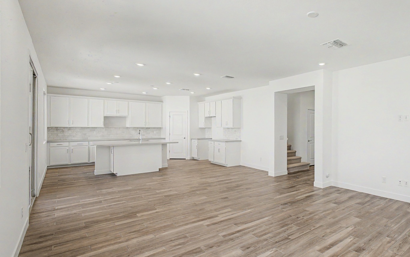 Open-concept kitchen and living area with white cabinets, light countertops, recessed lighting, and wood-style flooring. Stairs are visible in the background.