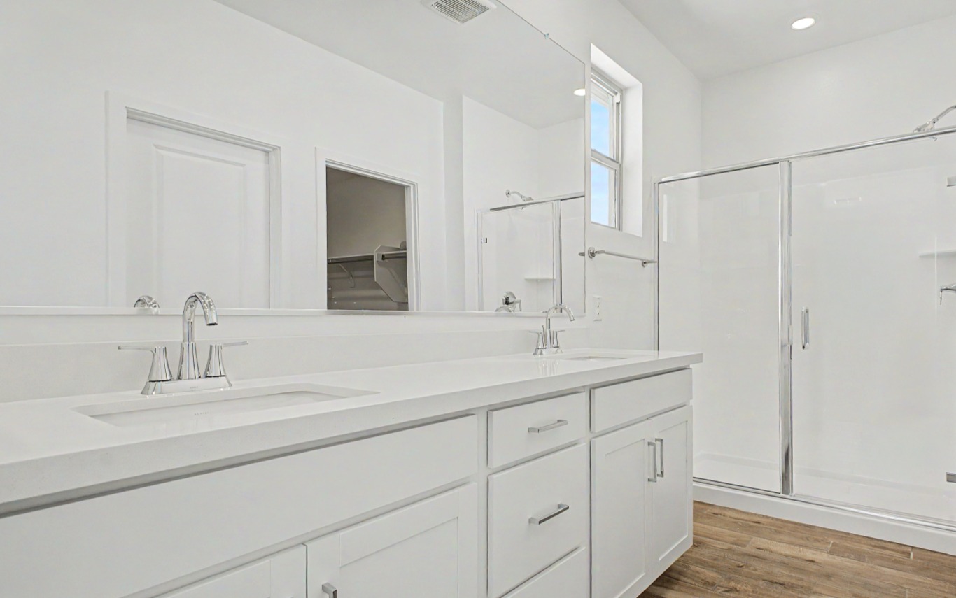 Modern bathroom with white double-sink vanity, large mirror, glass shower enclosure, and wood-style flooring. Bright lighting and a minimalist design are featured throughout the space.