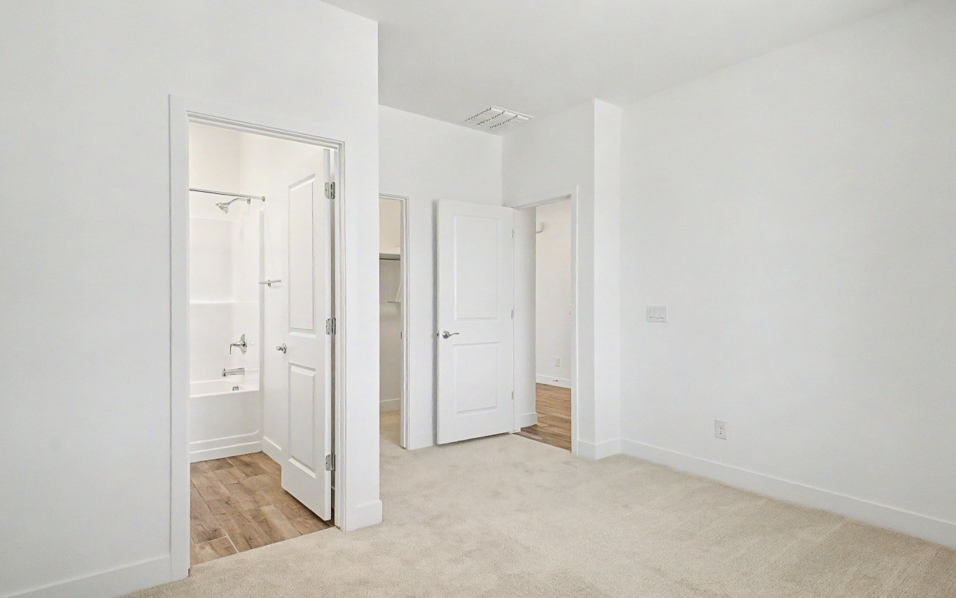 Empty, white-walled bedroom with carpet flooring, two open doors, and an ensuite bathroom featuring a bathtub with tiled floor.
