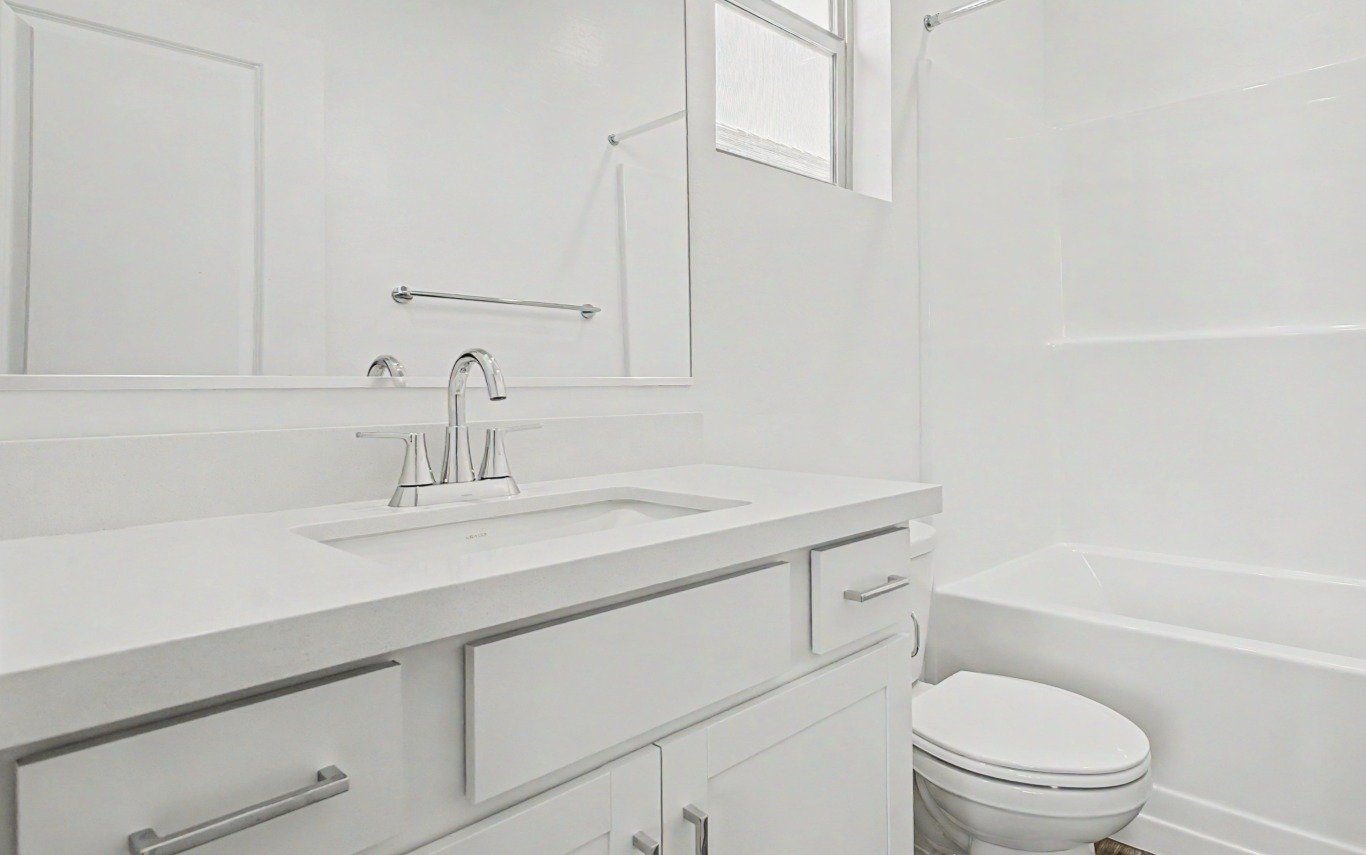 Modern white bathroom with a vanity sink, chrome faucet, drawers, toilet, towel rack, and a bathtub with shower. Large mirror and frosted window above the sink.