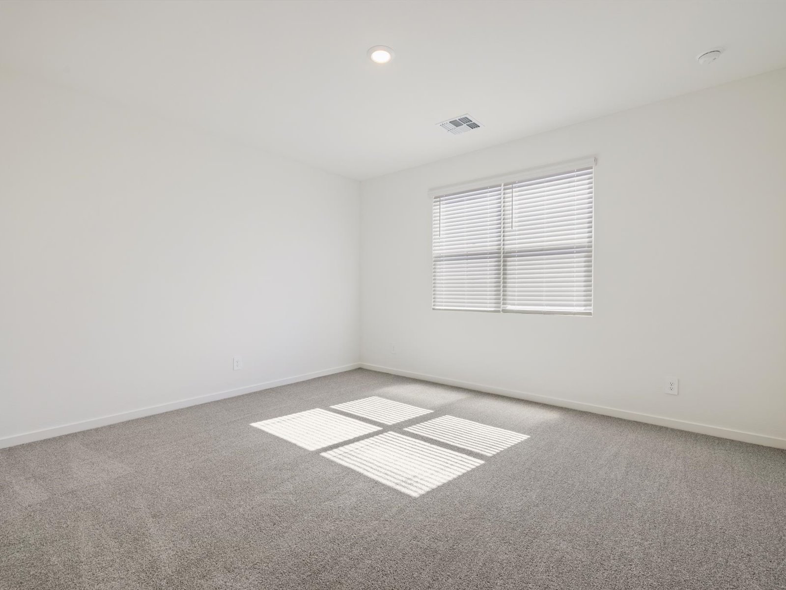 Empty room with light gray carpet, white walls, and a window with blinds letting in sunlight. Ceiling light fixture is on.