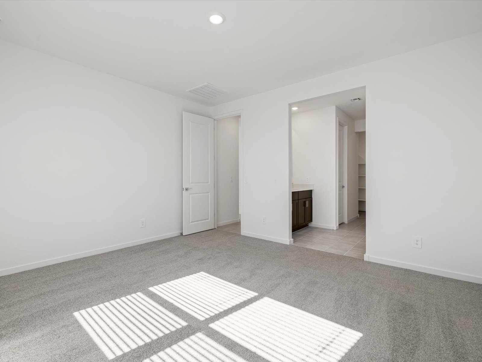Empty bedroom with light gray carpet, white walls, a door leading to another room, and sunlight streaming through window blinds onto the floor.