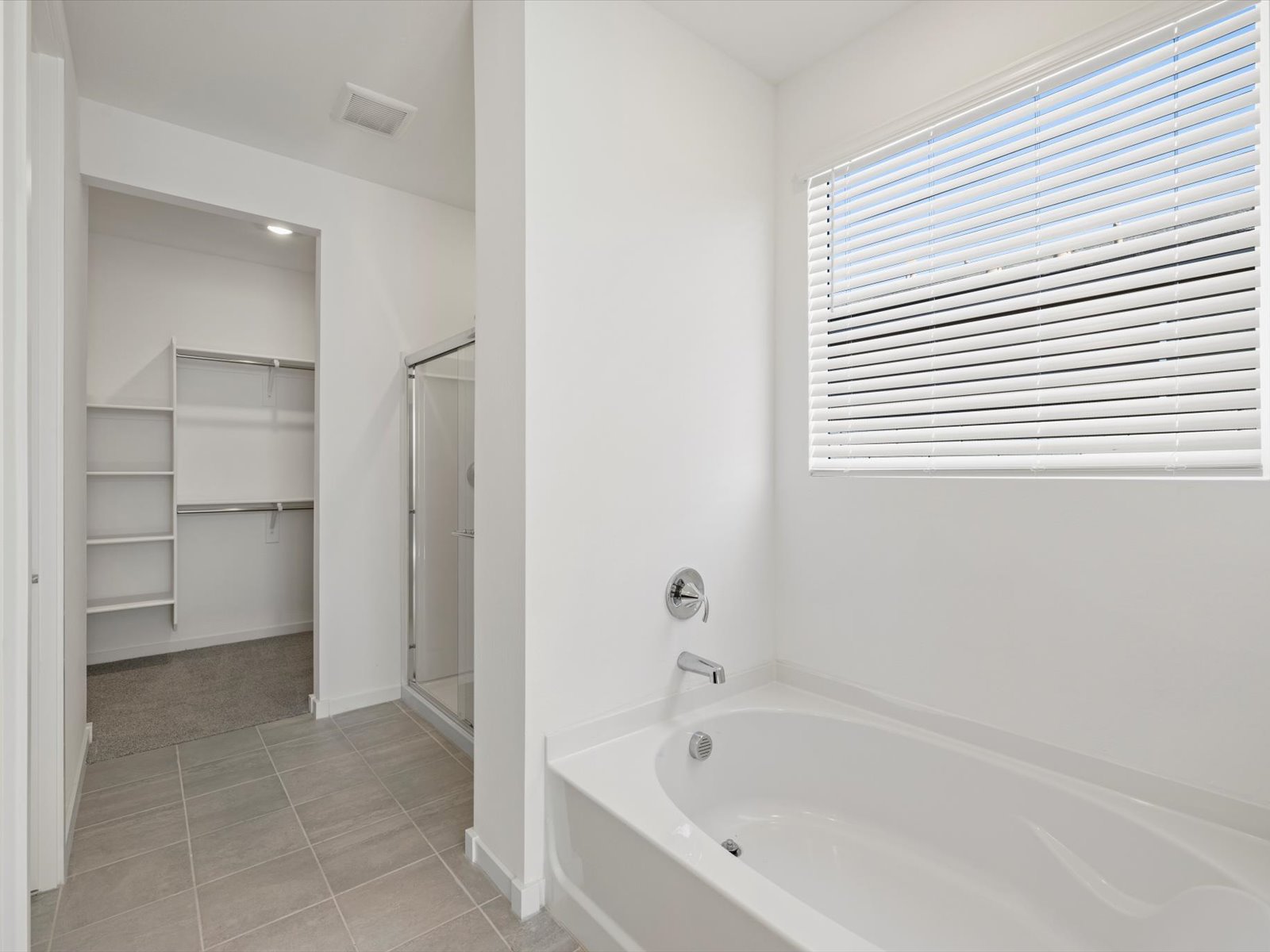 Modern bathroom with a white bathtub, glass shower, large window with blinds, tile floor, and an adjacent walk-in closet with shelves and hanging rods.
