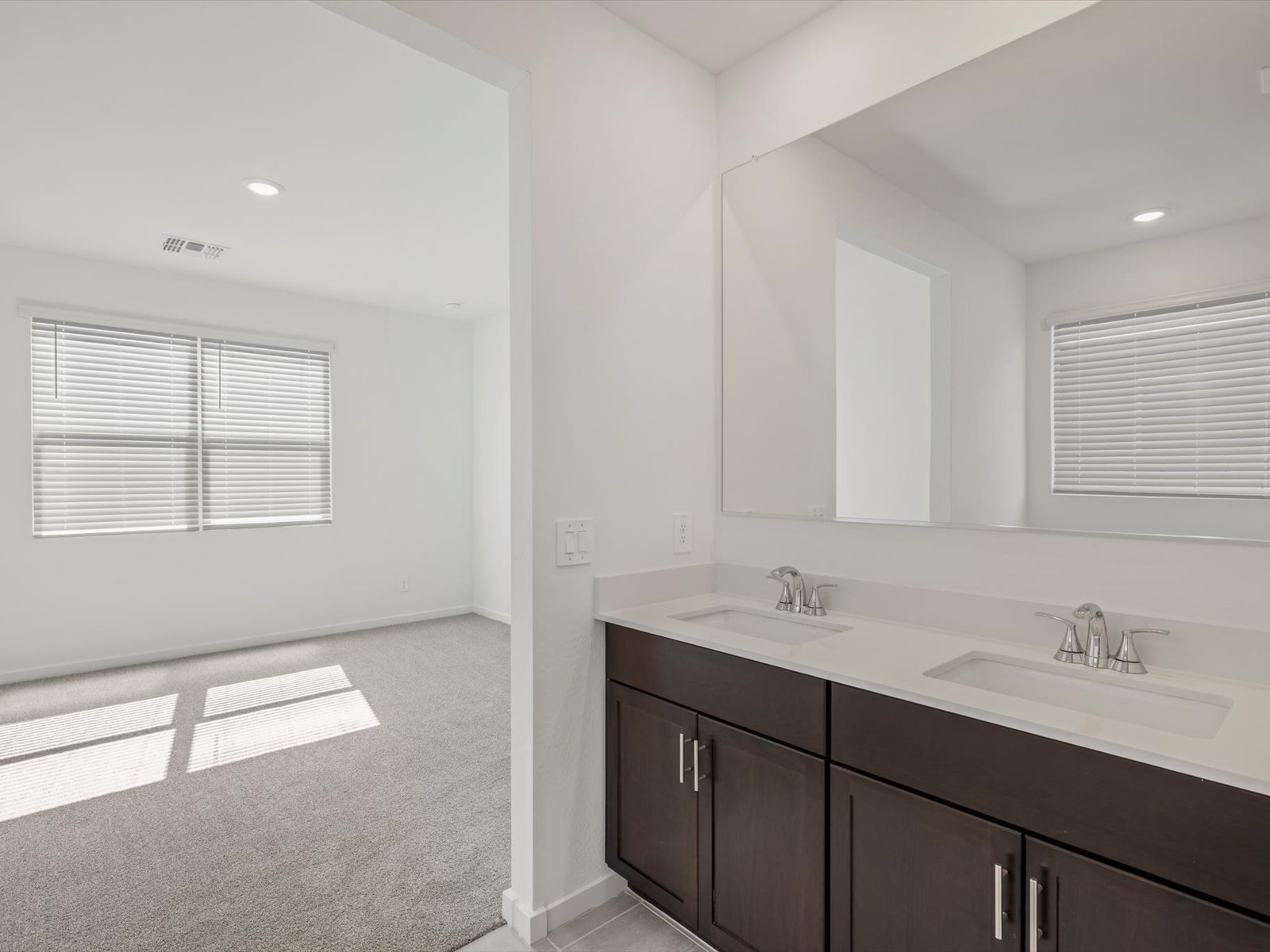 View of a bathroom with a double sink vanity and a large mirror, adjacent to a carpeted bedroom with two windows letting in natural light.
