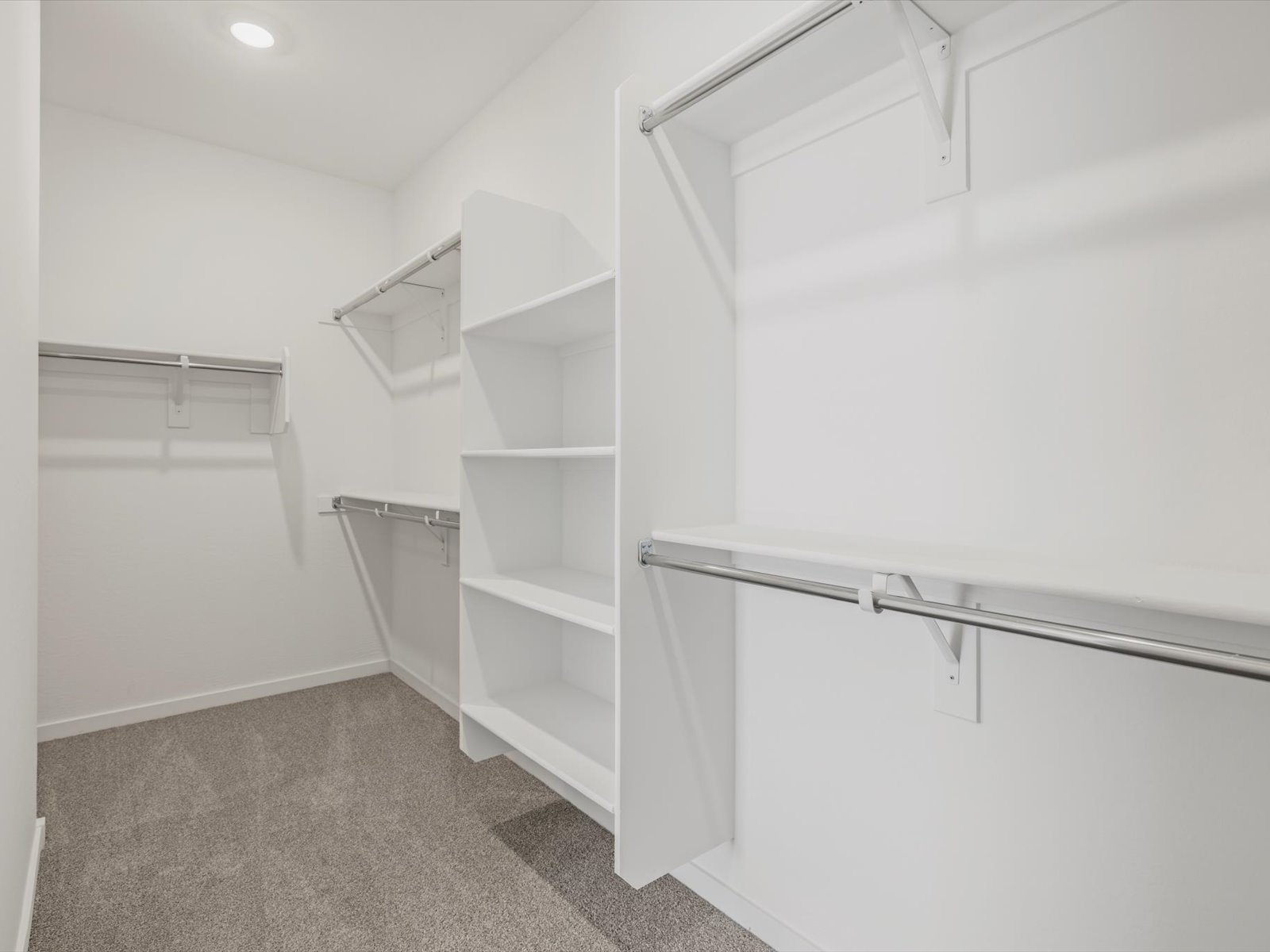 Empty walk-in closet with white shelves, multiple hanging rods, and beige carpet flooring under bright ceiling light.