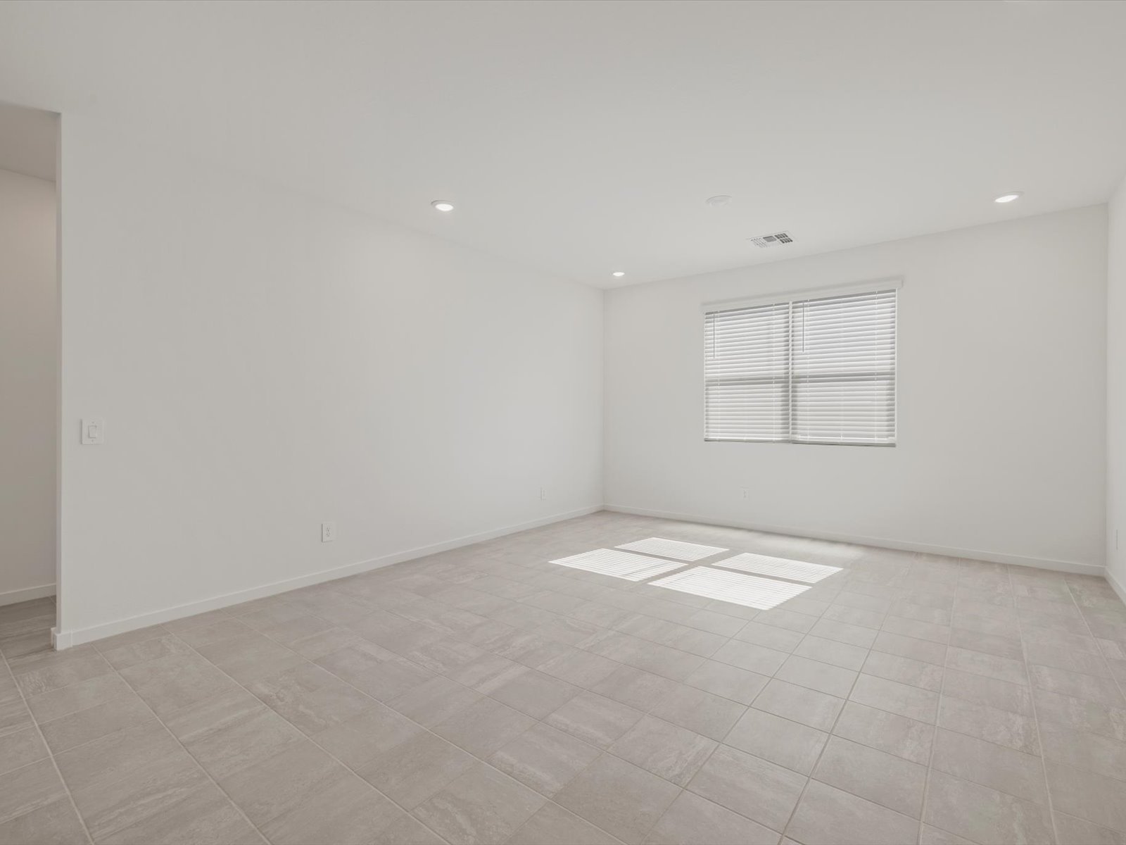 Empty room with white walls, large window with blinds, beige tiled floor, and recessed ceiling lights. Natural light streams in from the window.