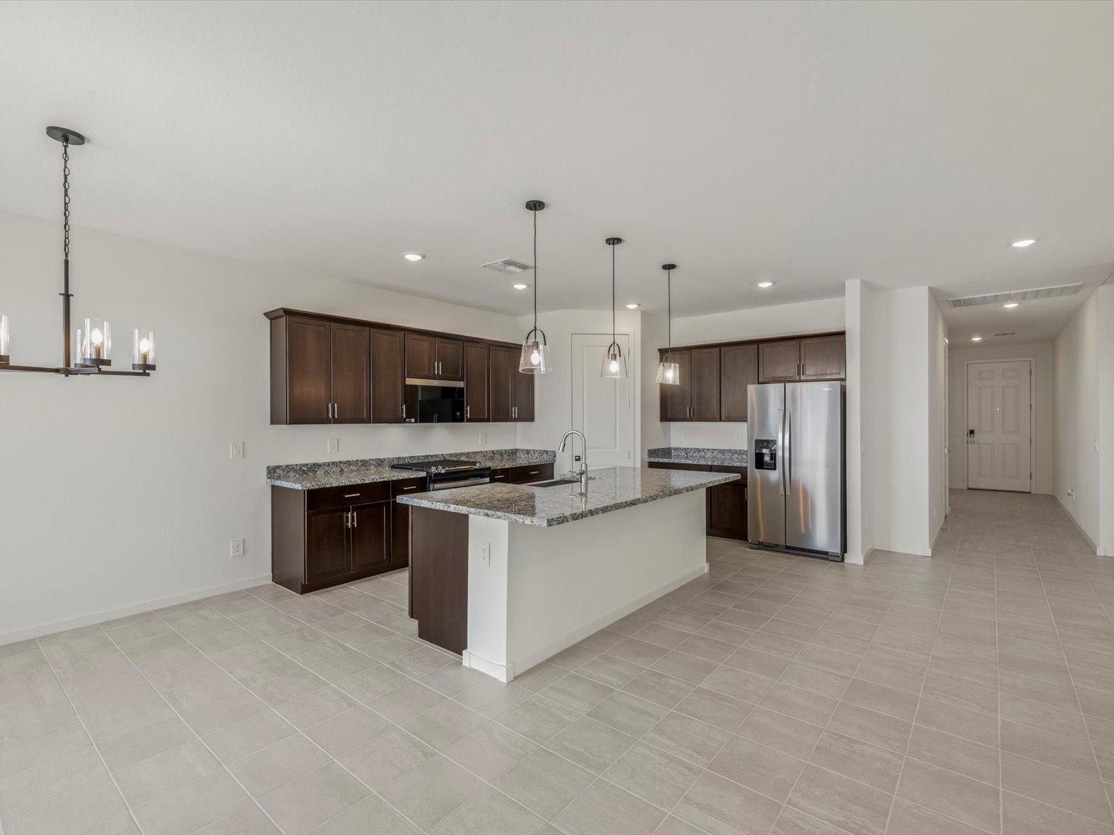 Modern kitchen with dark wood cabinets, stainless steel appliances, granite countertops, pendant lighting, and tile flooring, adjacent to a dining area and hallway with white walls.