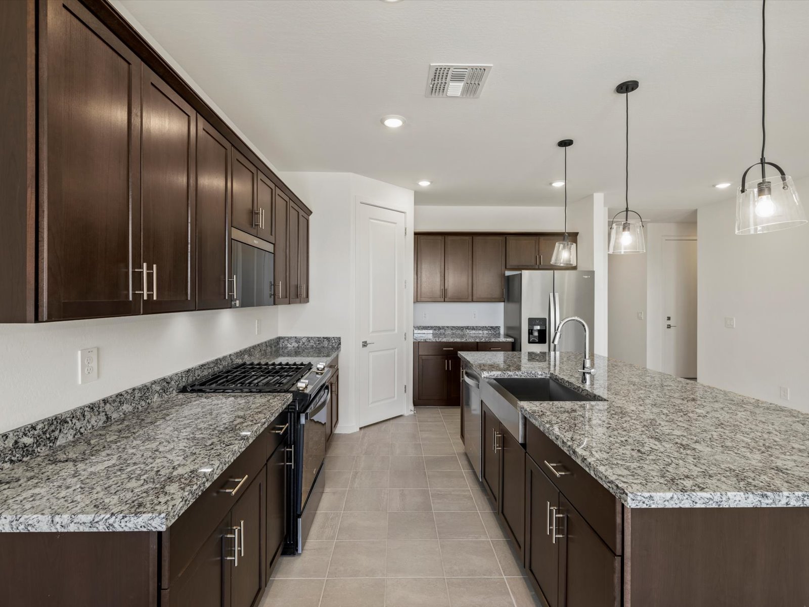 Modern kitchen with dark wood cabinets, granite countertops, stainless steel appliances, tile flooring, pendant lights, and a double-basin sink. The room is well-lit and spacious.