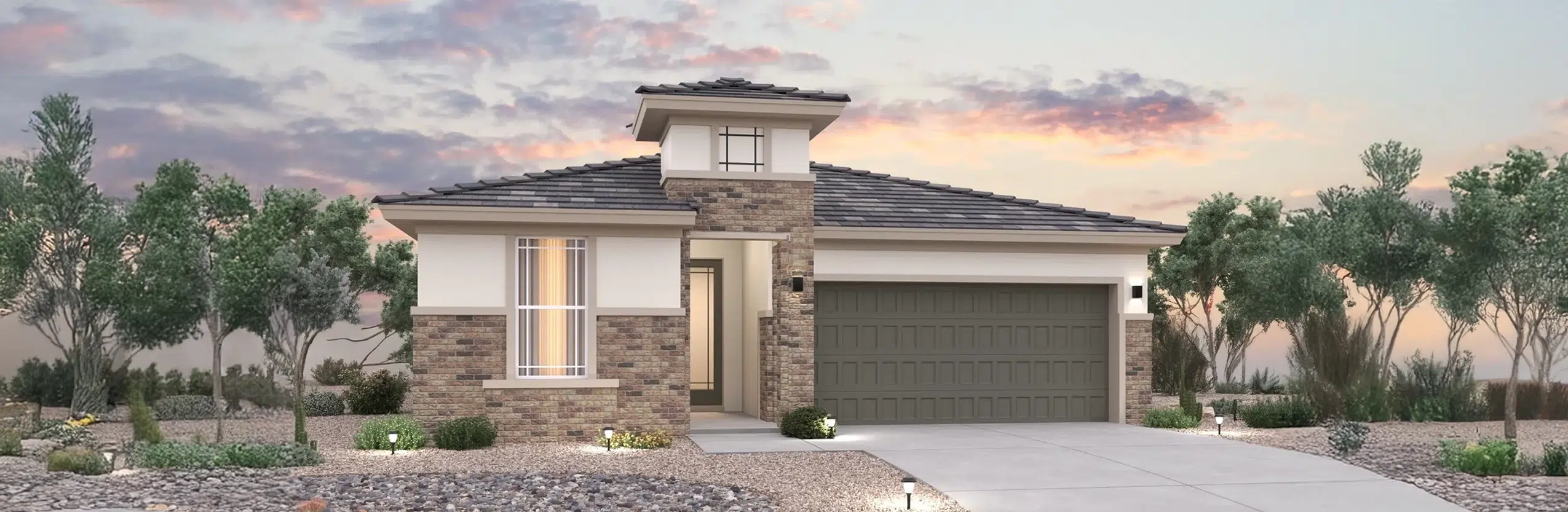 A modern single-story house with brick and stucco exterior, a two-car garage, desert landscaping, and outdoor lighting at sunset.