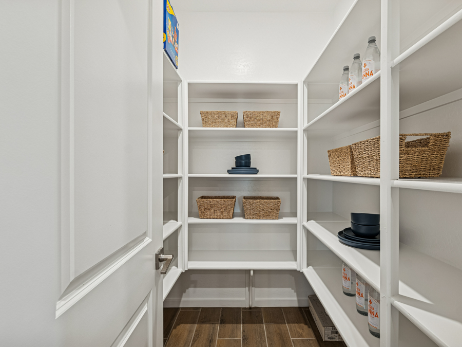 Walk-in pantry with white shelves, wicker baskets, bottled drinks, dark blue plates and bowls, and wooden floor.