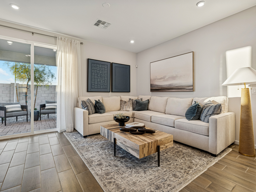 Modern living room with a beige sectional sofa, wooden coffee table, area rug, floor lamp, wall art, and sliding glass doors leading to a patio with outdoor seating.