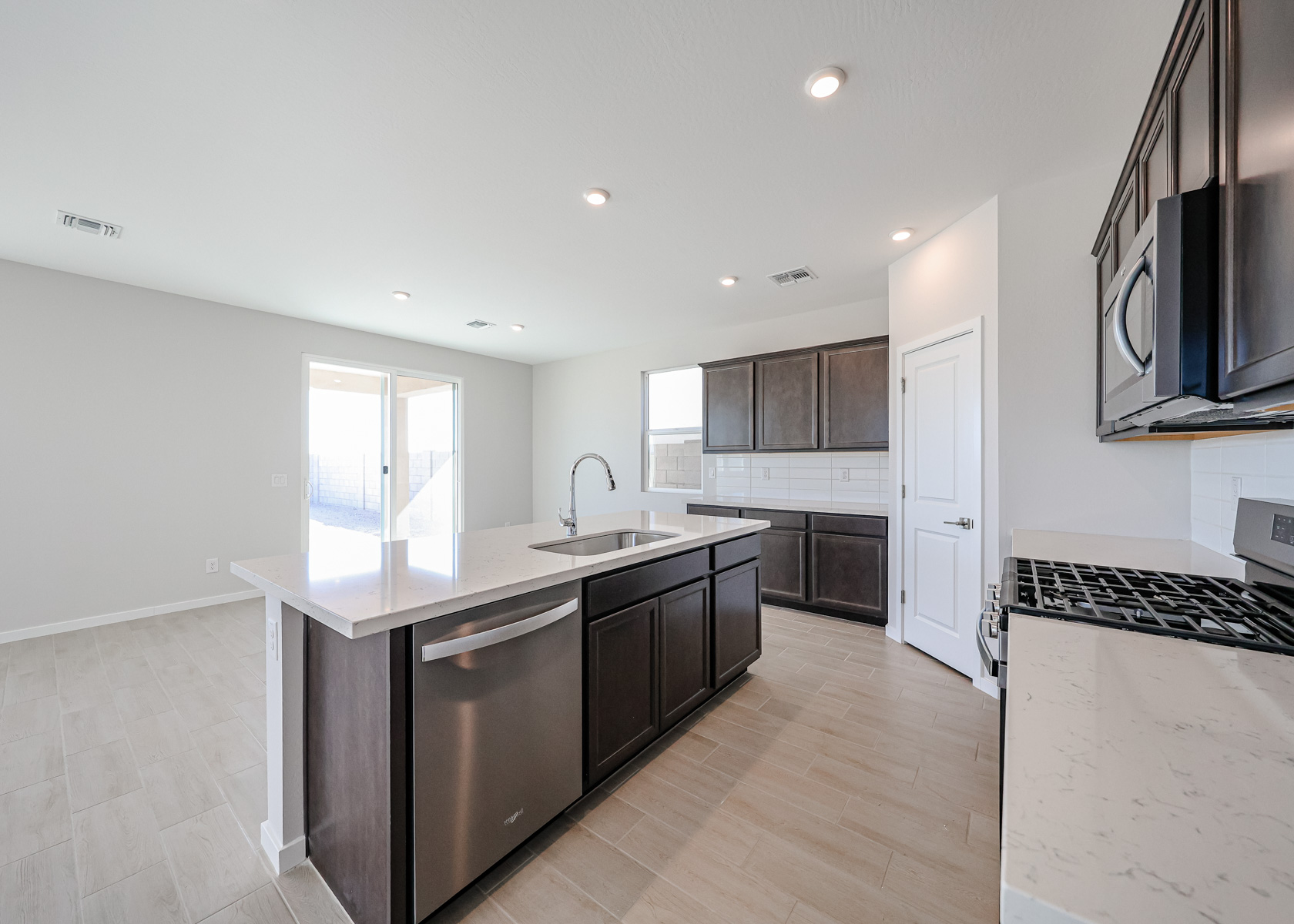 Modern kitchen with dark wood cabinets, stainless steel appliances, a center island with sink, light countertops, tile flooring, and ample natural light from large windows and glass door.