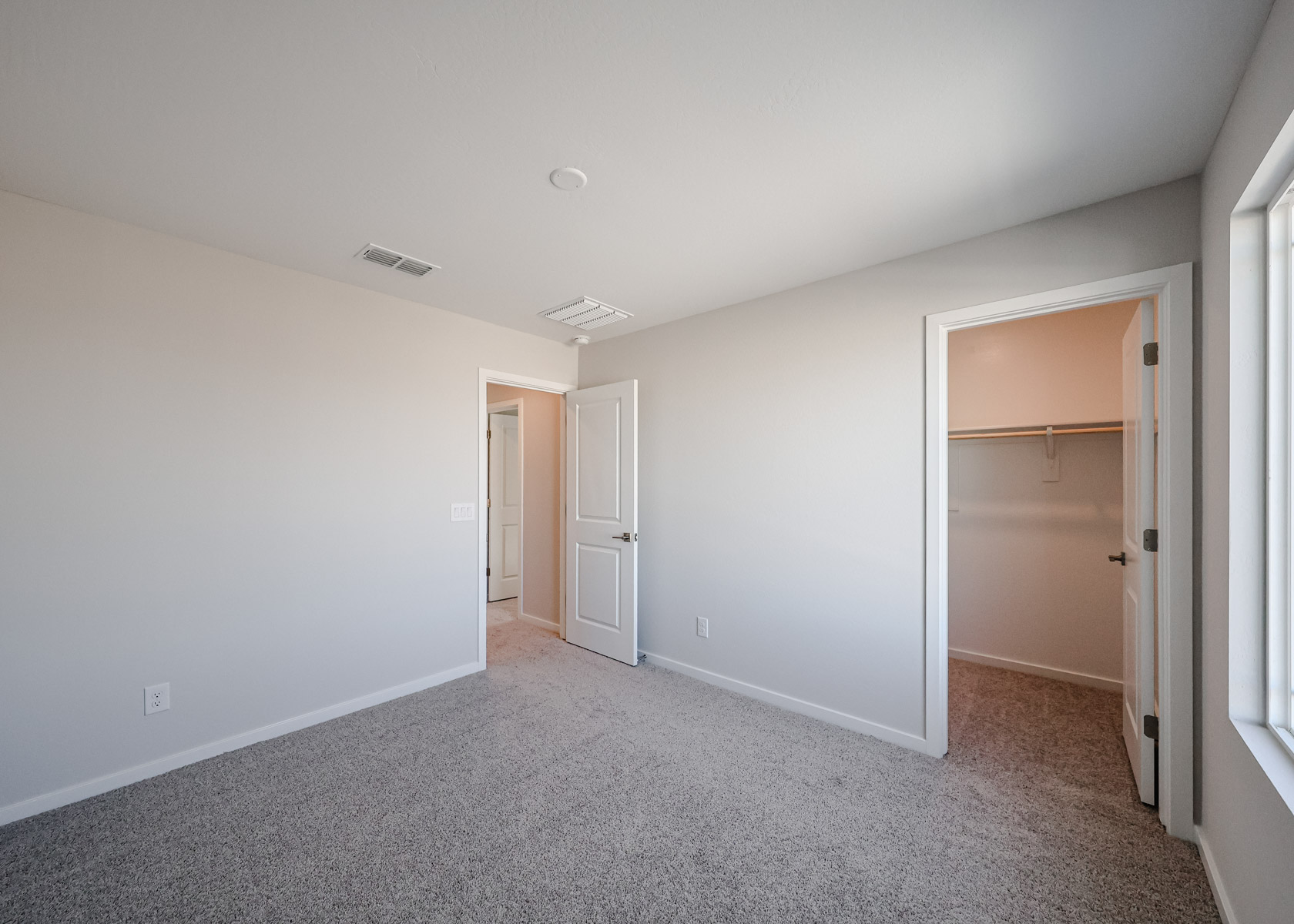 Empty bedroom with beige carpet, white walls, an open door to a hallway, and an open door leading to a walk-in closet. Sunlight enters through a window on the right.