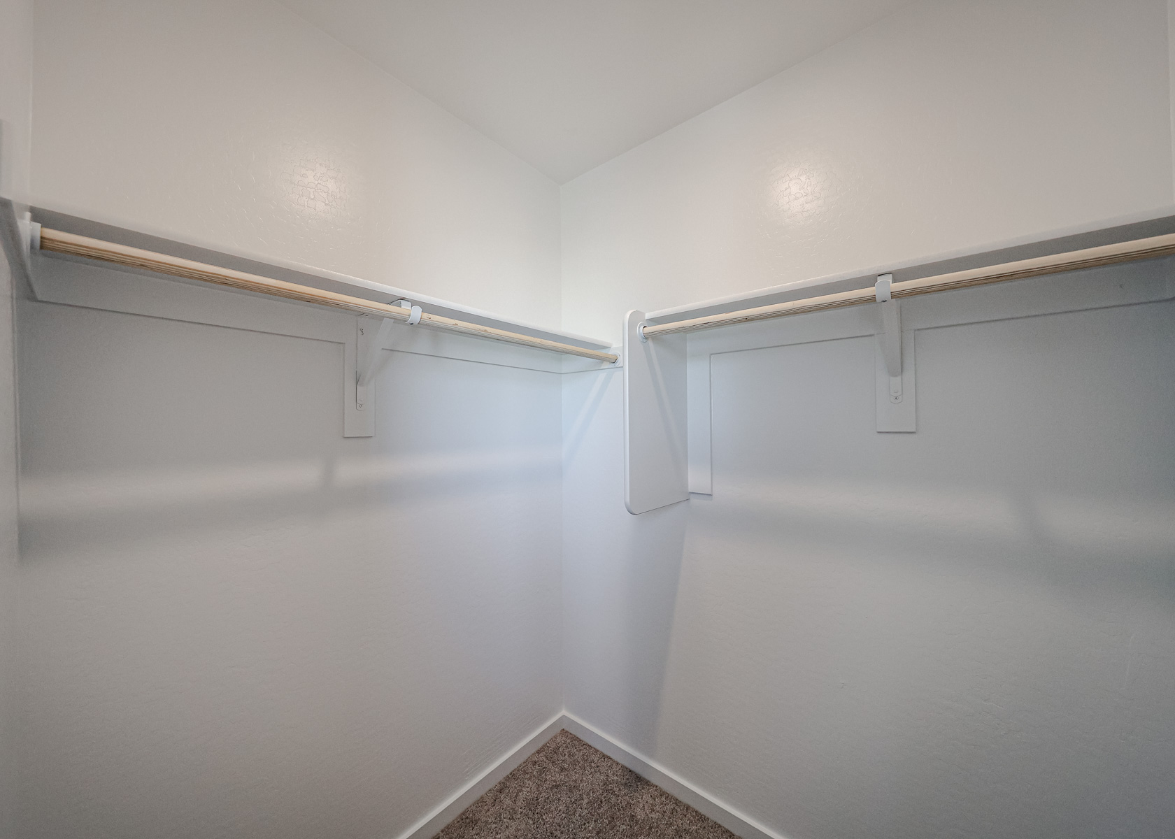 Empty walk-in closet with white walls, two wooden hanging rods, and a beige carpeted floor.