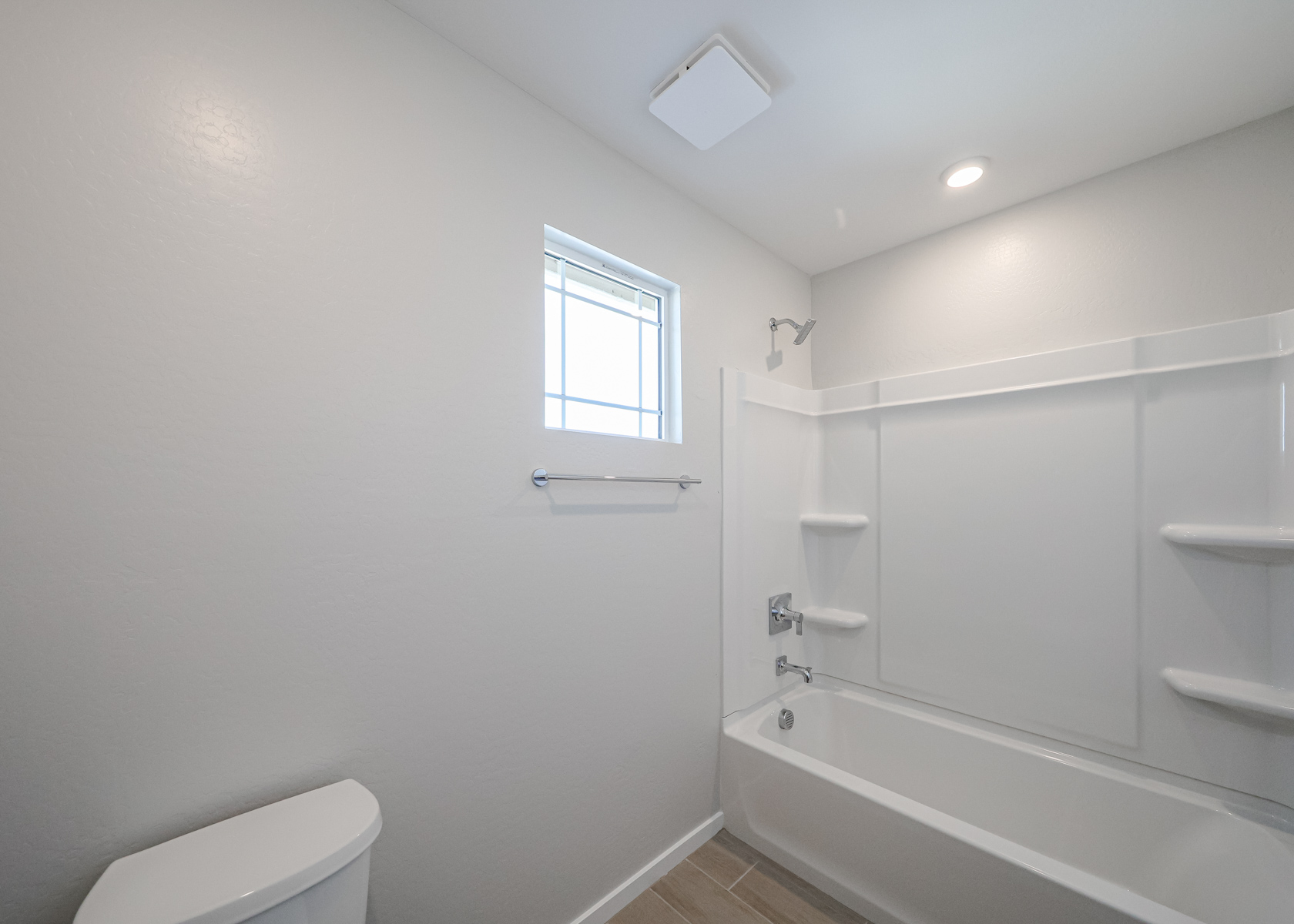 A clean, white bathroom with a bathtub-shower combo, a small window, and a toilet. The walls are plain, and natural light enters through the window.