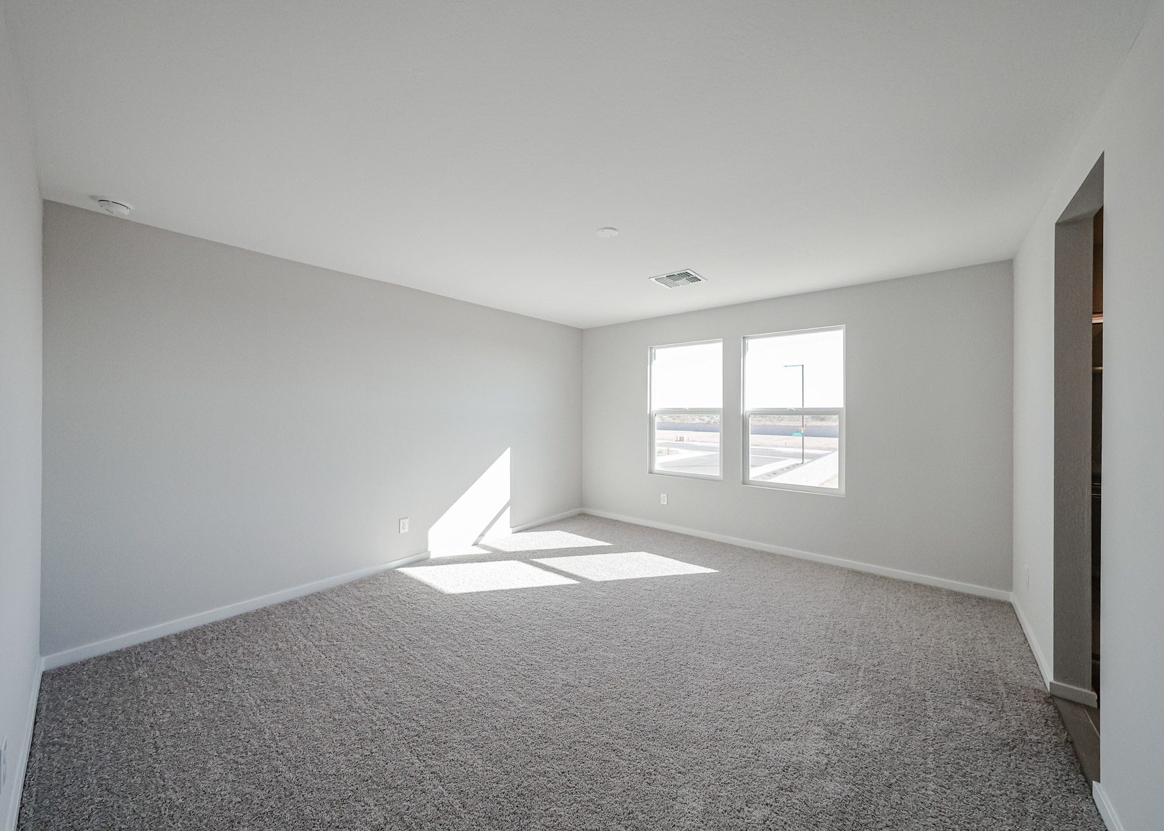 Empty, carpeted room with light gray walls, two windows letting in natural light, and a view of the outdoors.