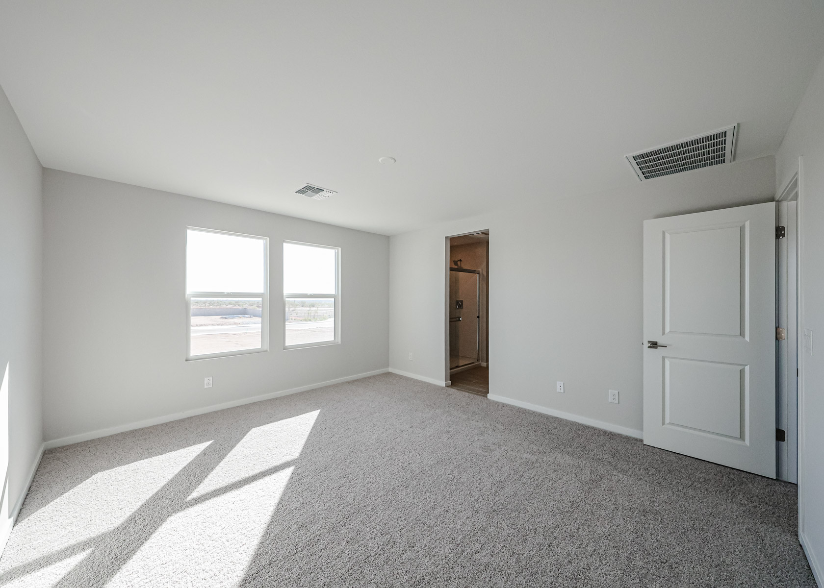 Empty, carpeted bedroom with two large windows, white walls, and an open door leading to a bathroom. Sunlight streams in from the windows, casting shadows on the floor.