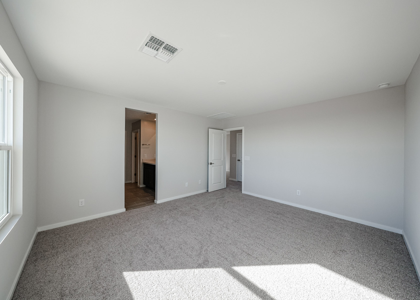 Empty, unfurnished bedroom with gray carpet, beige walls, two windows, and an open doorway leading to a bathroom or hallway area.