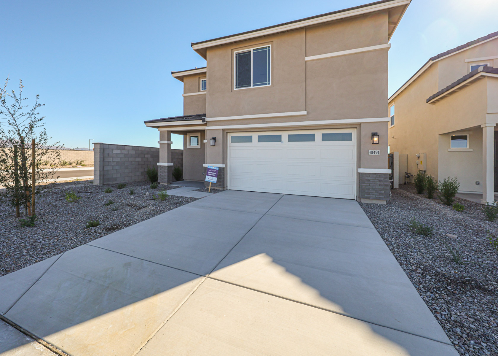 Two-story beige house with a two-car garage, concrete driveway, and minimal desert landscaping in front.