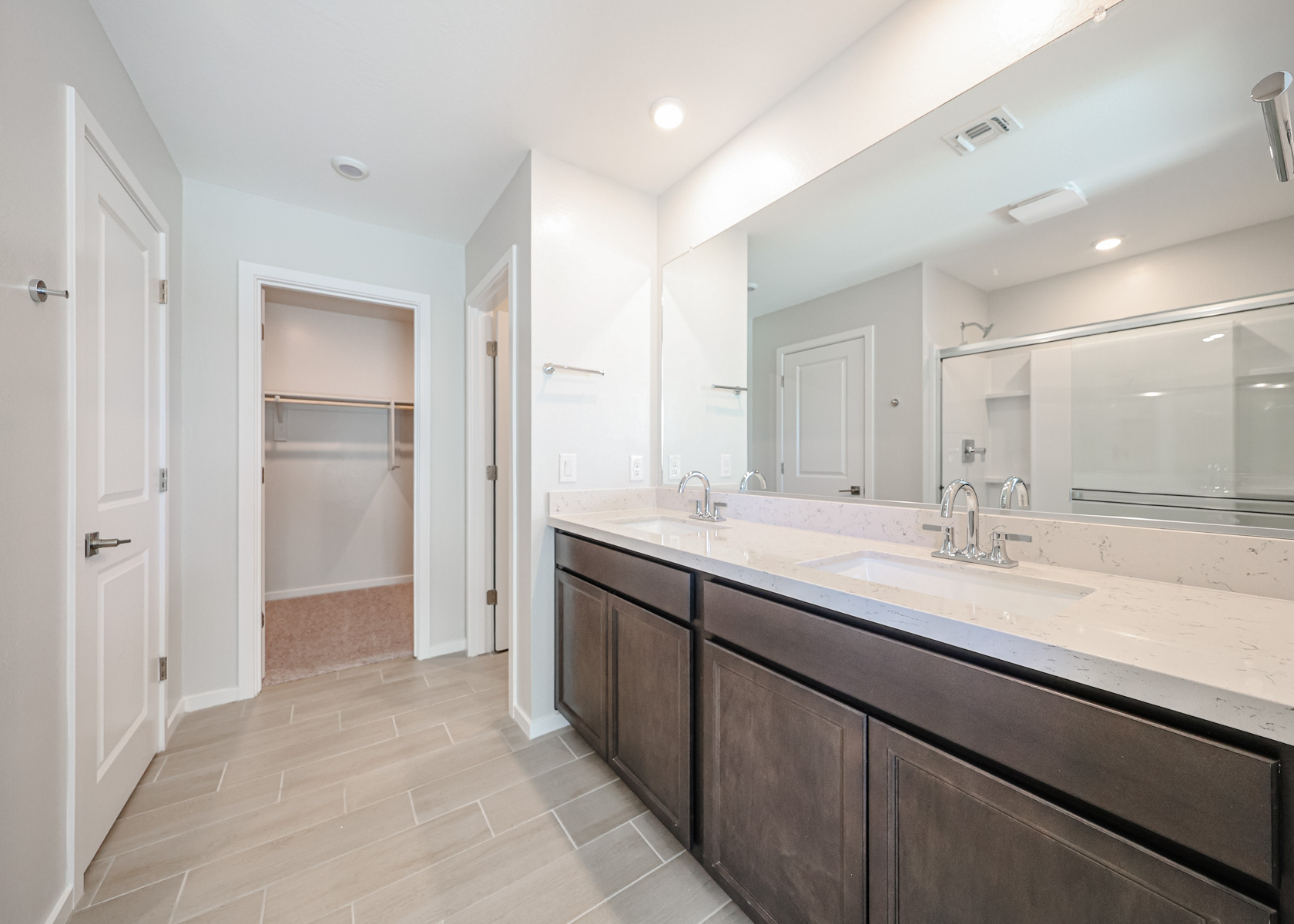 Modern bathroom with double sinks, large mirror, glass shower, and walk-in closet with open shelving. Light gray floor tiles and white walls.