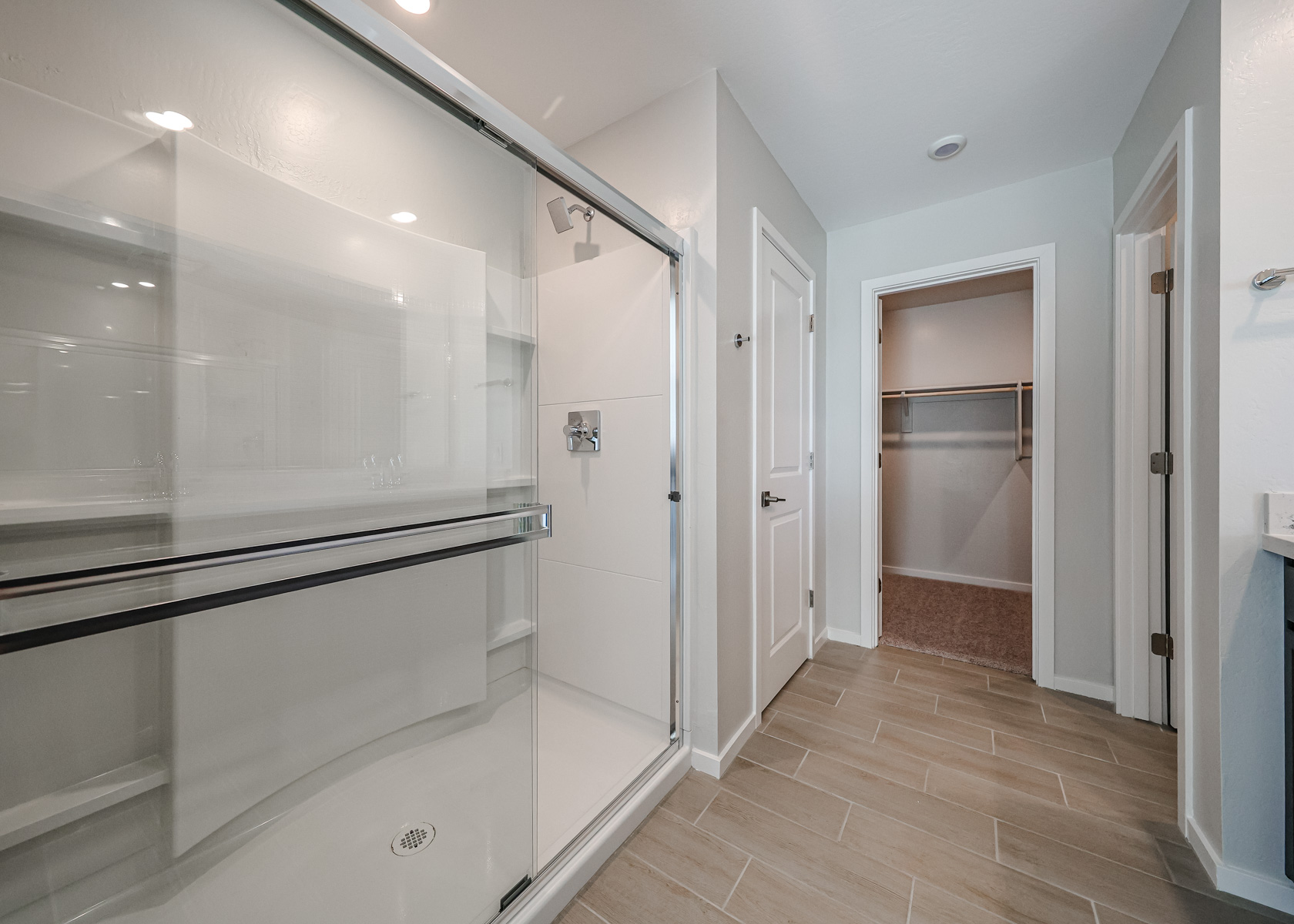 Modern bathroom with a large glass-enclosed shower, beige tile flooring, two white doors, and an open walk-in closet with shelving.
