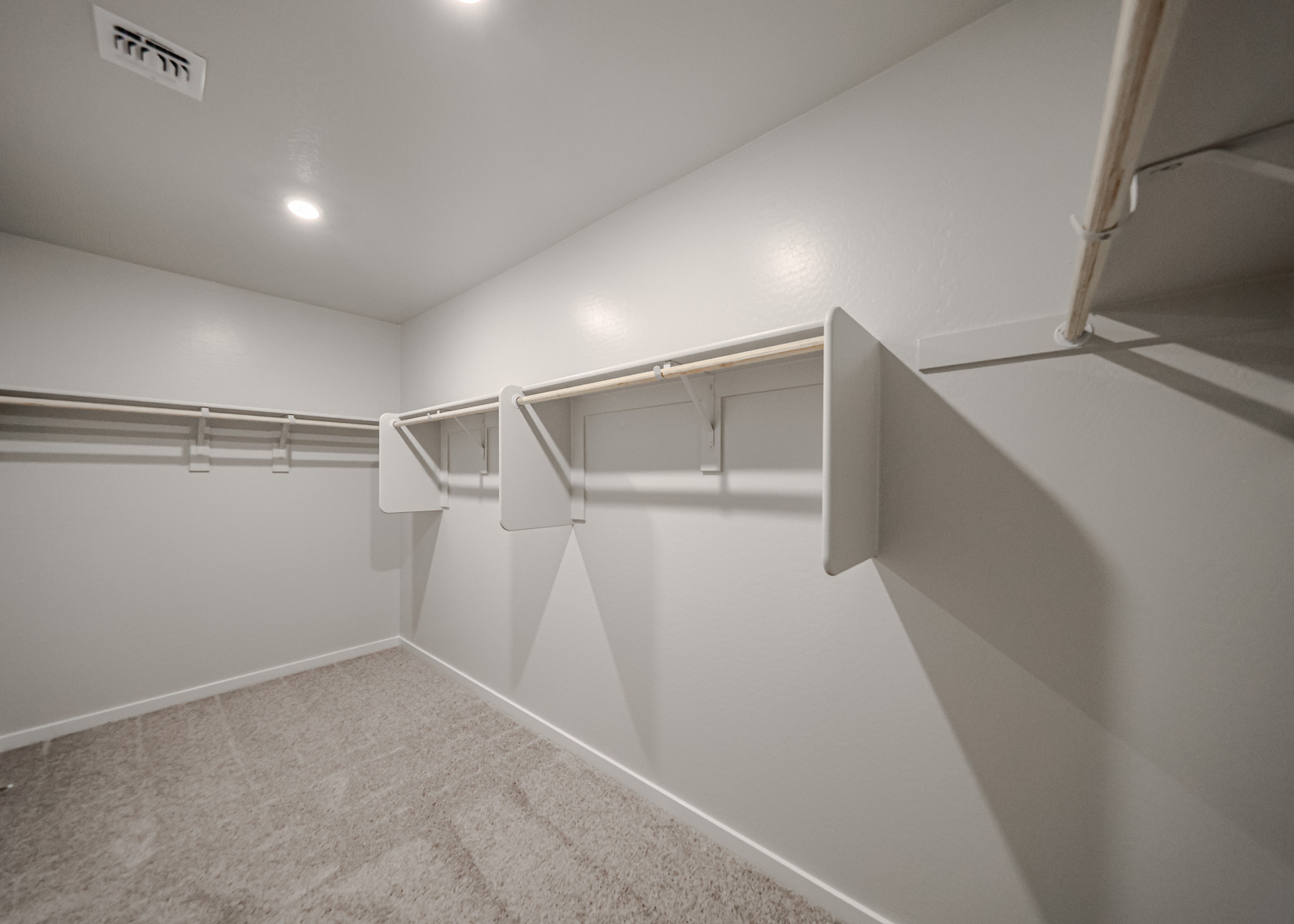 Empty walk-in closet with beige carpet, white walls, and multiple wooden hanging rods and shelves on both sides. Ceiling vent and recessed lighting are visible.