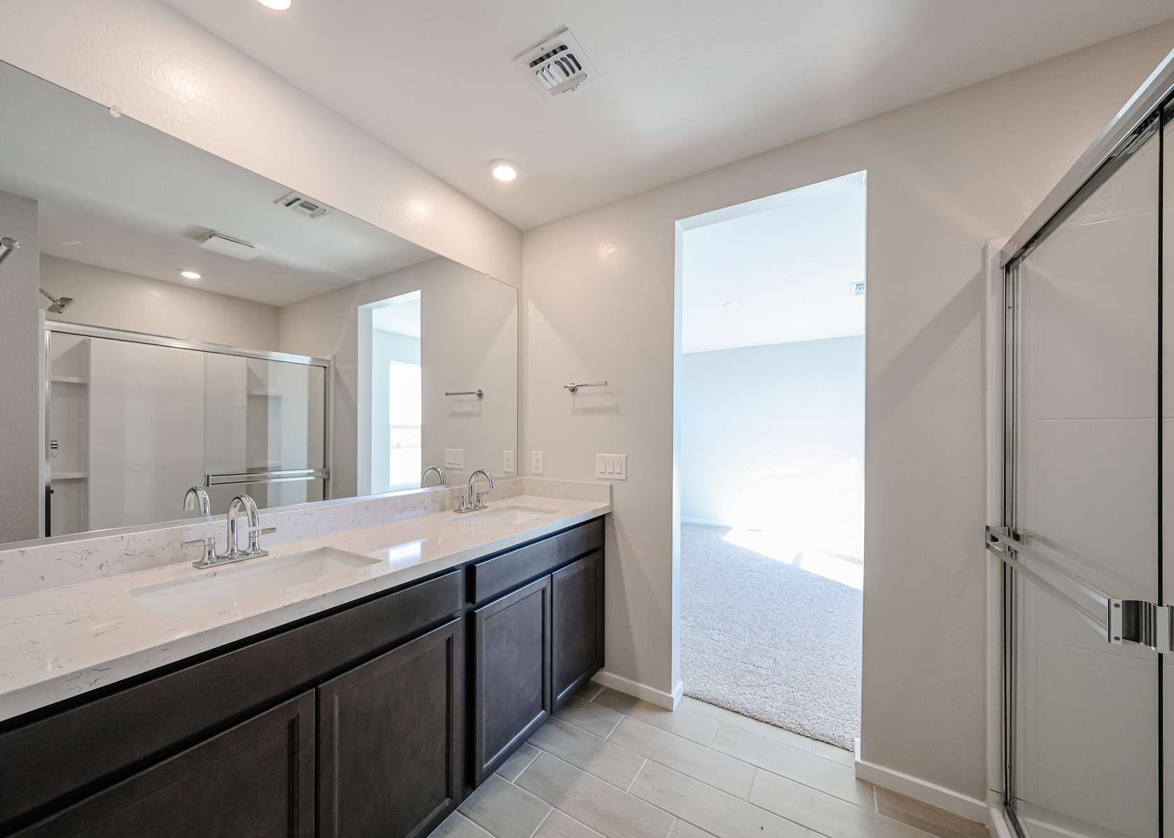 Modern bathroom with double sink vanity, large mirror, glass shower enclosure, and tiled floor, leading to a carpeted room with natural light.