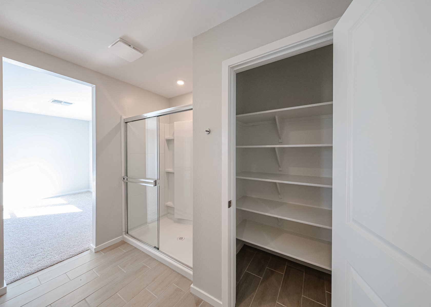 A bathroom with a glass-enclosed shower, beige tile flooring, and an open closet with white shelves and a brown floor.