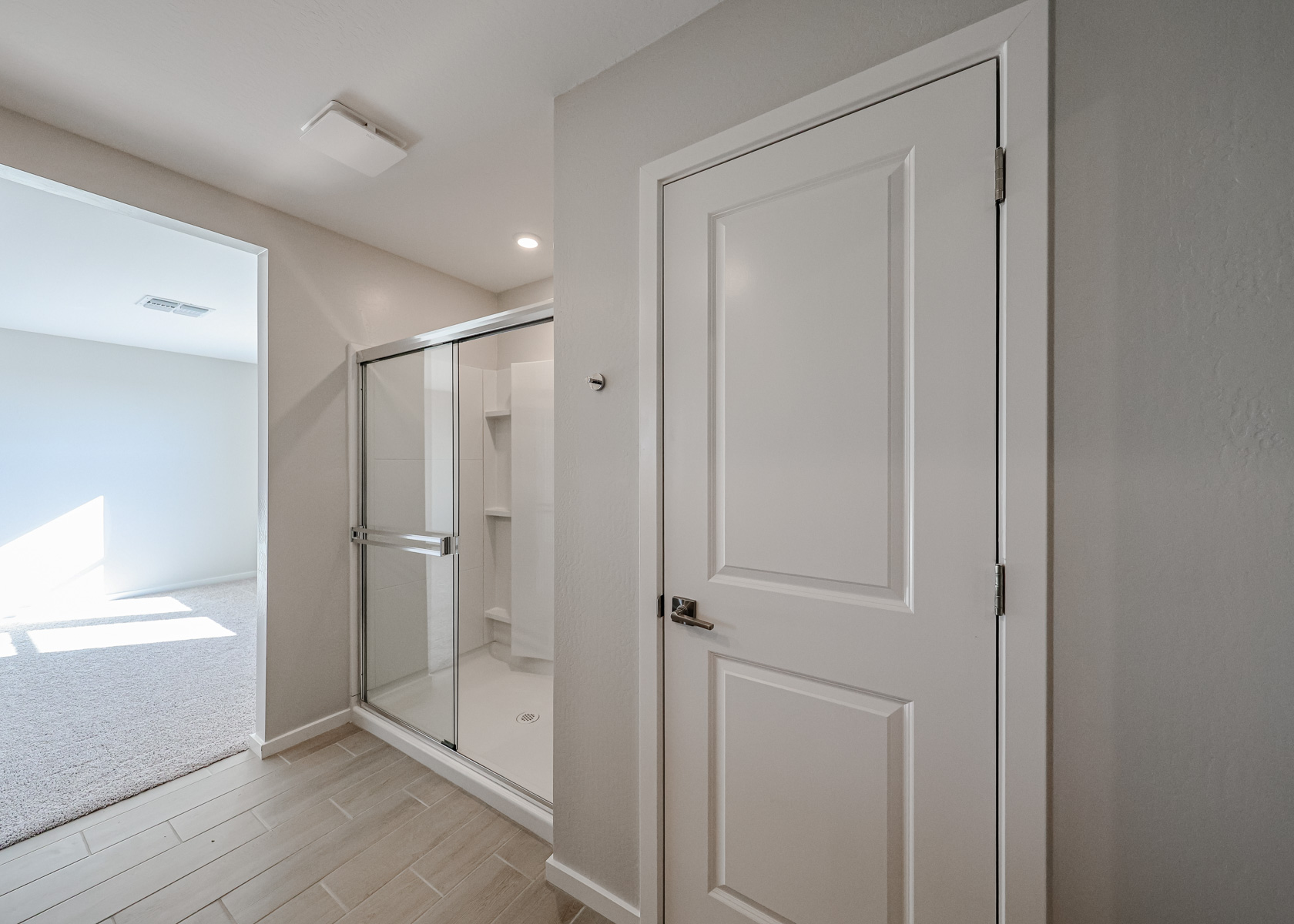 A bathroom with a white door, a shower with sliding glass doors, tiled flooring, and light gray walls. A carpeted room is visible through the open doorway.