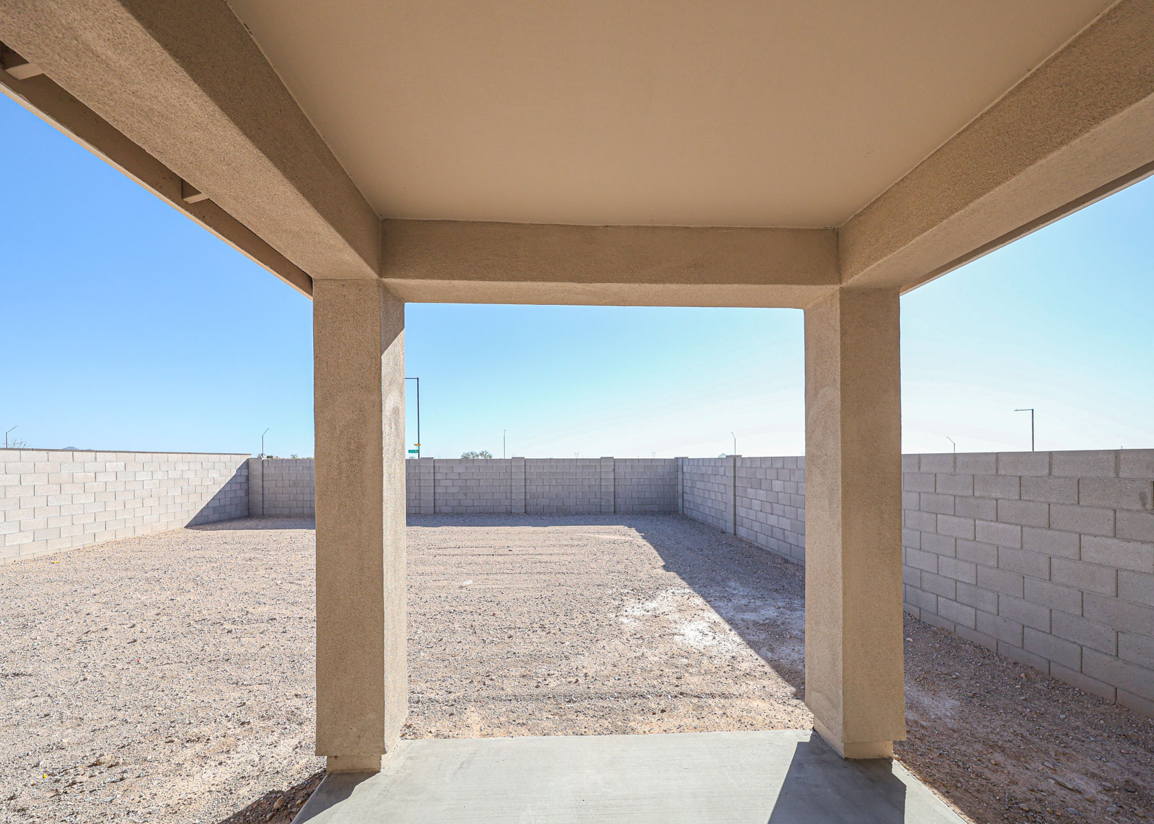 View from a covered patio looking out onto a bare, gravel backyard enclosed by a tall cinder block wall under a clear blue sky.
