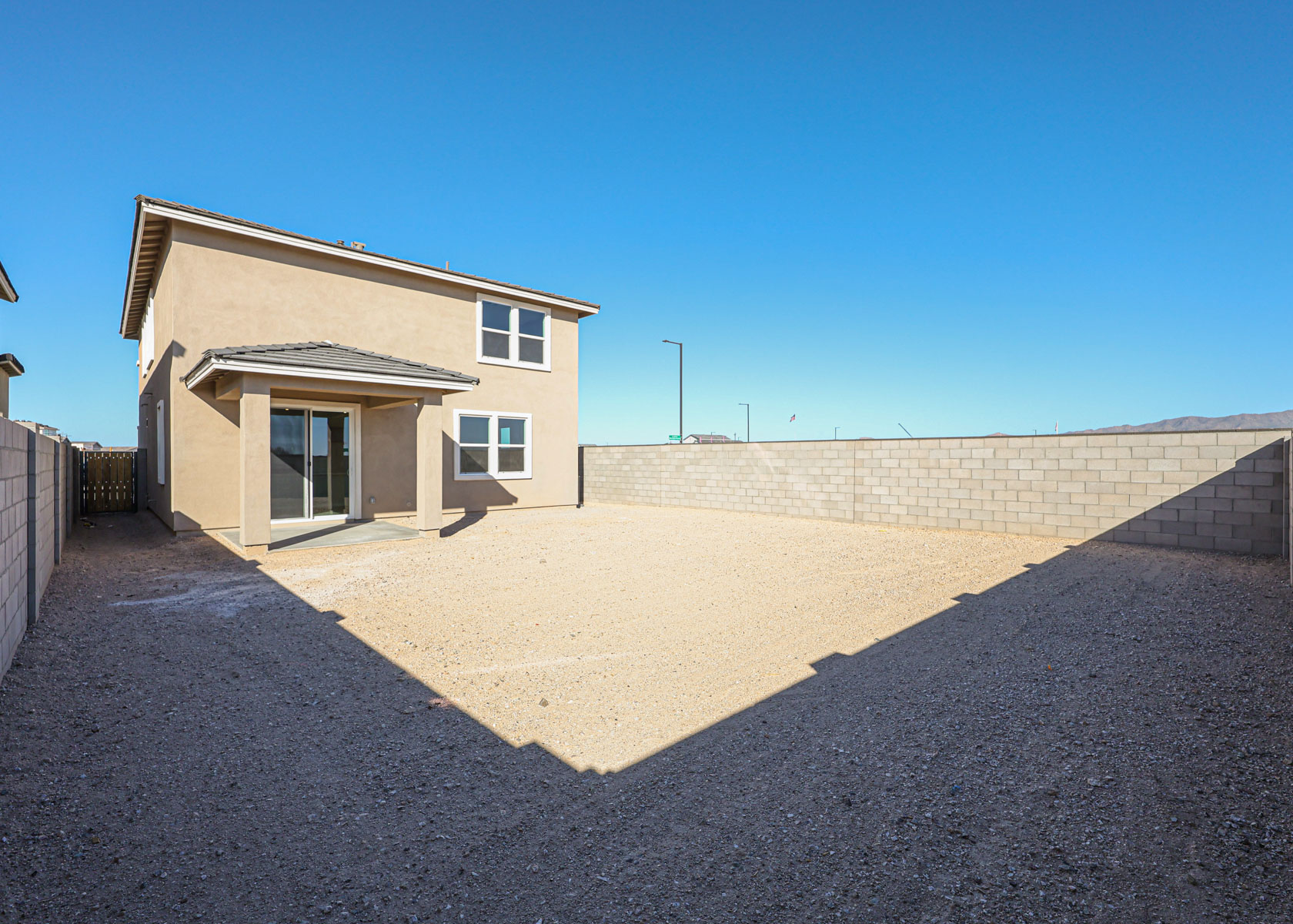 Two-story beige house with a sliding glass door opens to a large, bare backyard enclosed by a high concrete block wall under a clear blue sky.