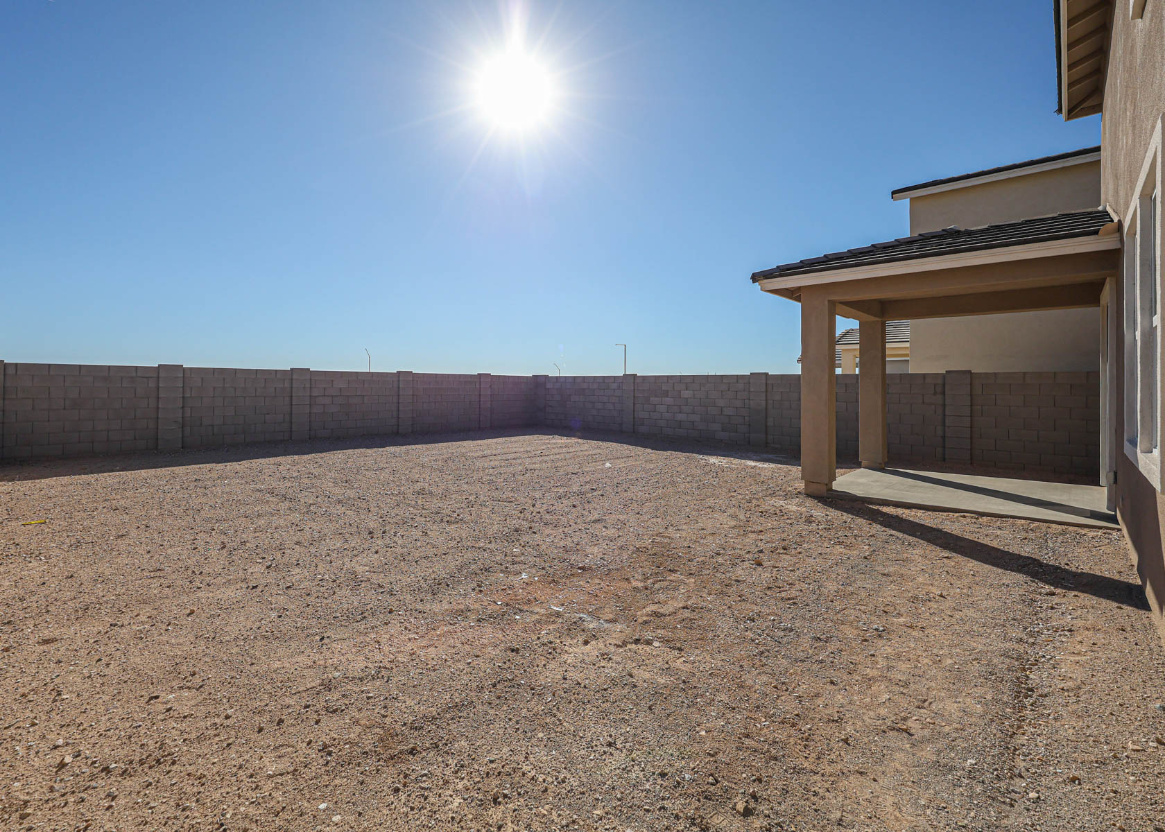 A fenced backyard with bare dirt ground, a high block wall, and a covered patio area beside a house under a clear, sunny sky.