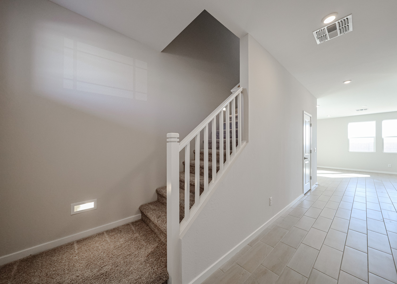 A modern, neutral-toned interior features carpeted stairs with a white railing and tiled flooring leading to a bright, open area.