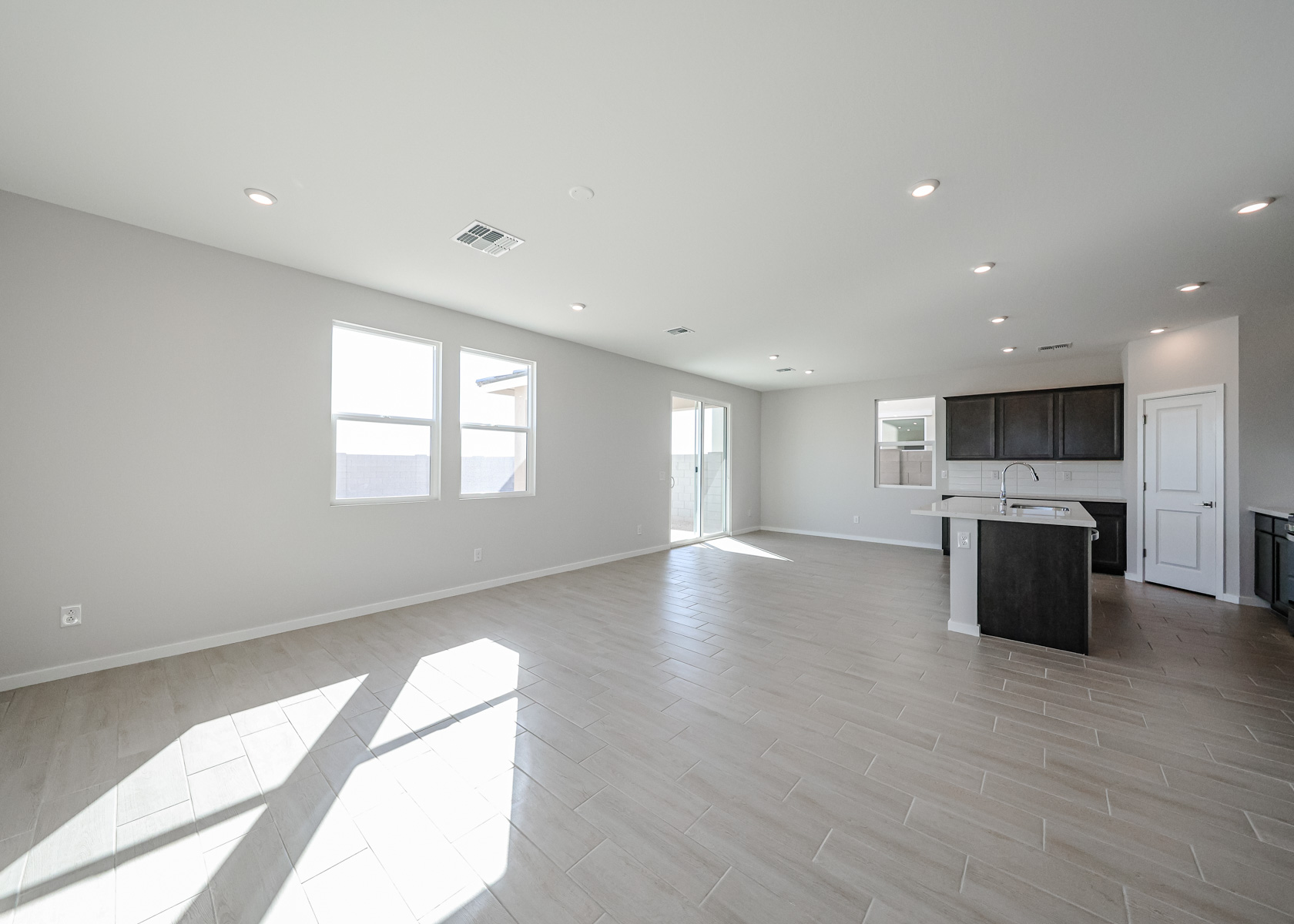 Spacious empty living area with light tile flooring, white walls, large windows, recessed lighting, and an open kitchen with dark cabinets and a center island.