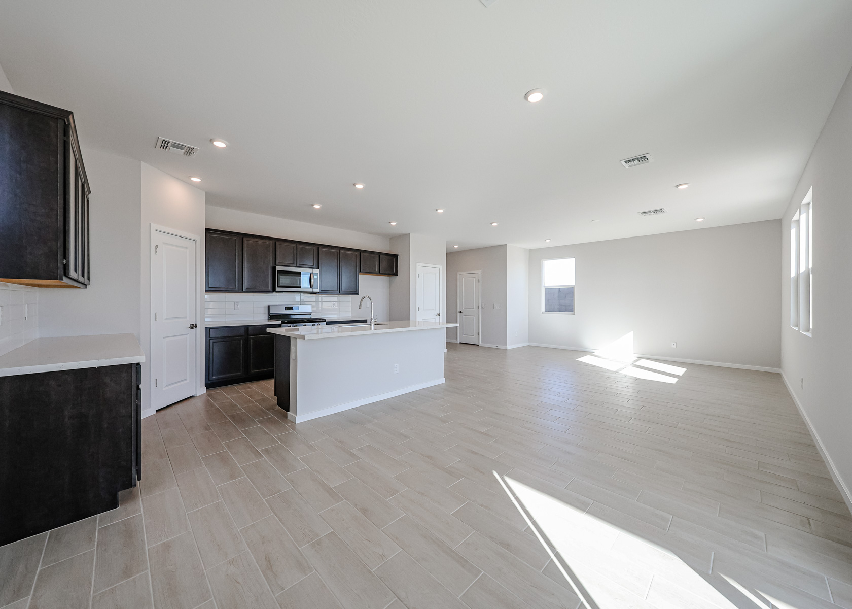 Spacious, unfurnished open-plan kitchen and living area with tiled floors, dark cabinets, white walls, and multiple windows letting in natural light.