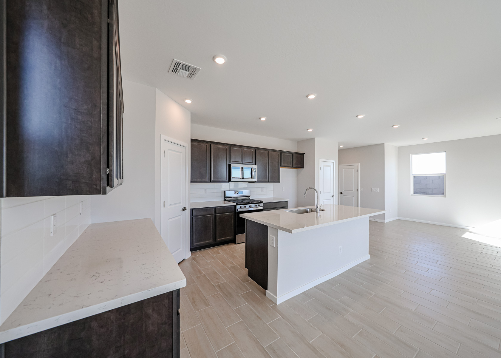Modern kitchen with dark cabinetry, stainless steel appliances, a central island with sink, light countertops, tile flooring, and recessed lighting, opening to a spacious living area.