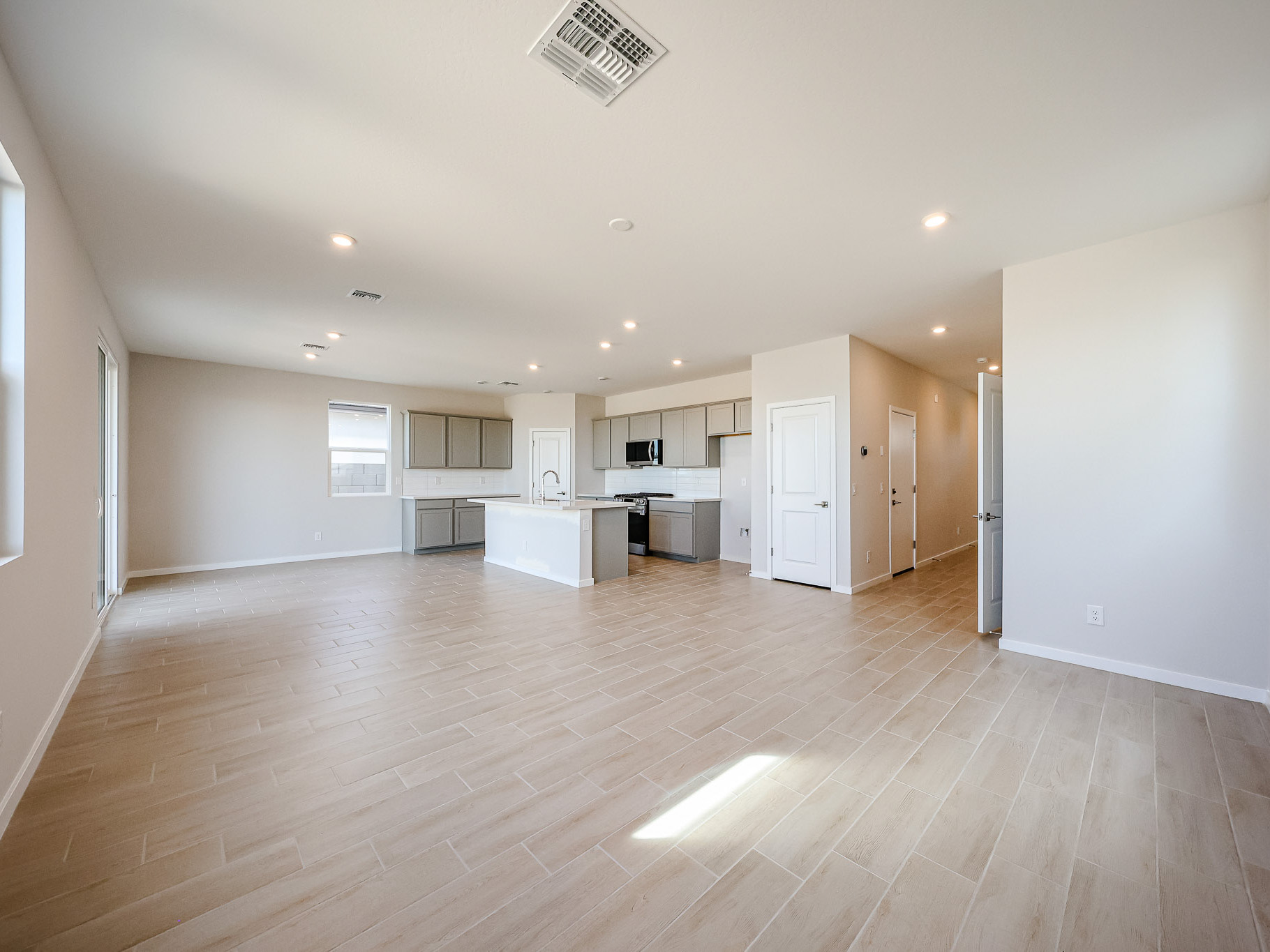 Spacious, empty open-concept living area with light wood-look tile flooring, recessed lighting, and a modern kitchen with white cabinets and stainless steel appliances.