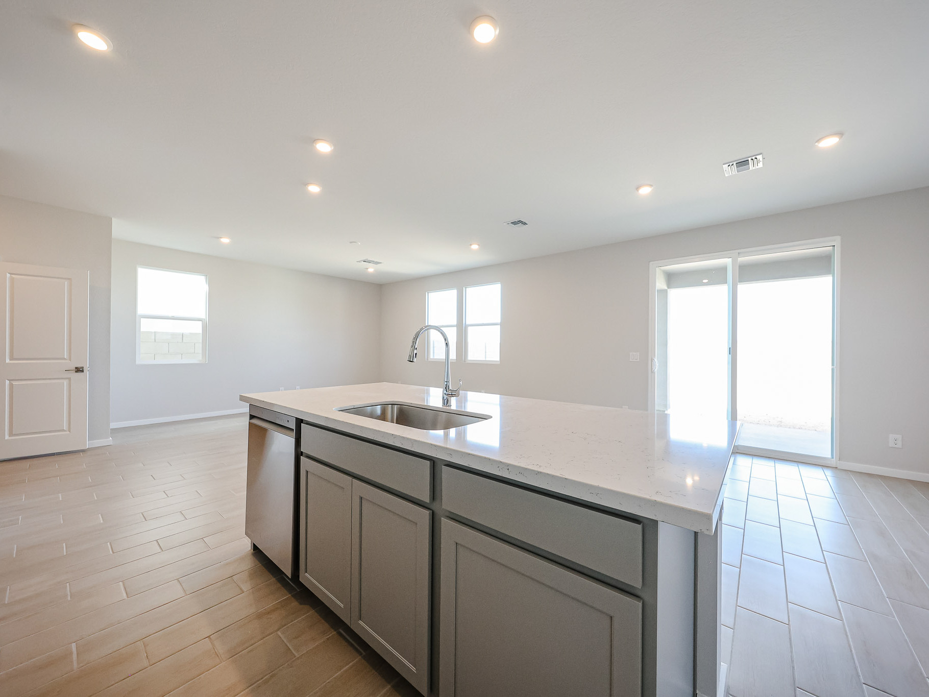 Bright, modern kitchen with a large island, stainless steel sink, light wood flooring, white walls, and sliding glass doors leading outside.