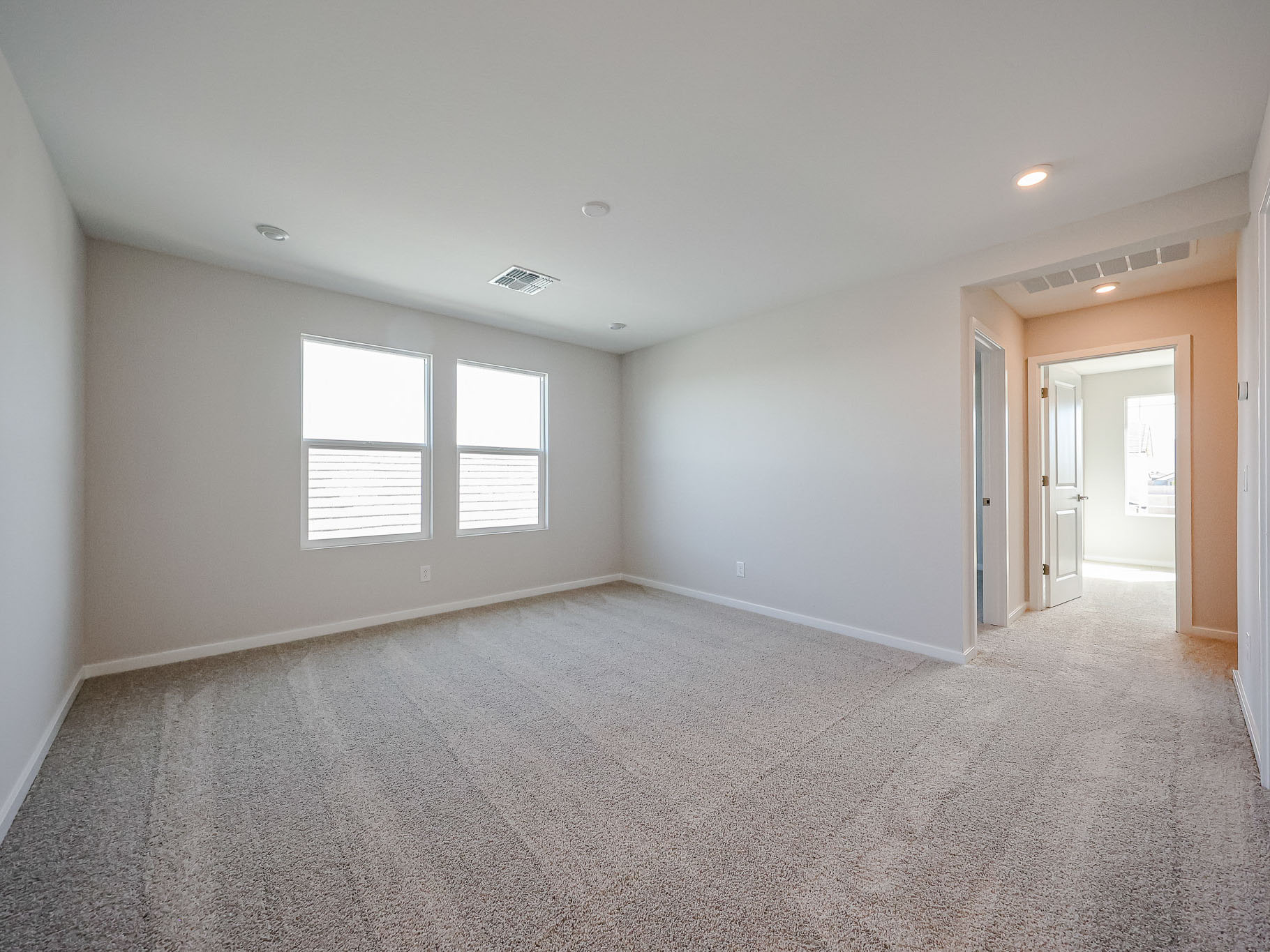 Empty, carpeted bedroom with two windows, neutral-colored walls, and an open doorway leading to a brightly lit hallway and bathroom.