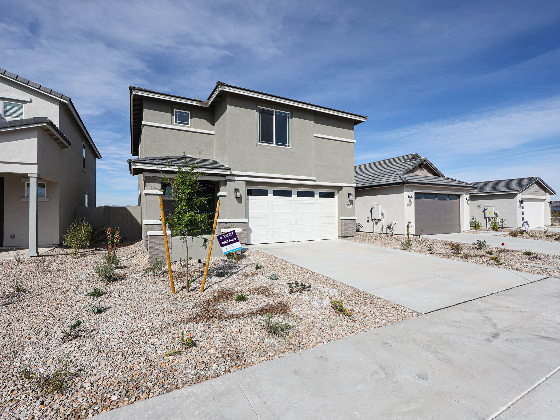 Two-story modern house with gray exterior, white garage door, and a "For Sale" sign in a xeriscaped front yard under a clear blue sky.