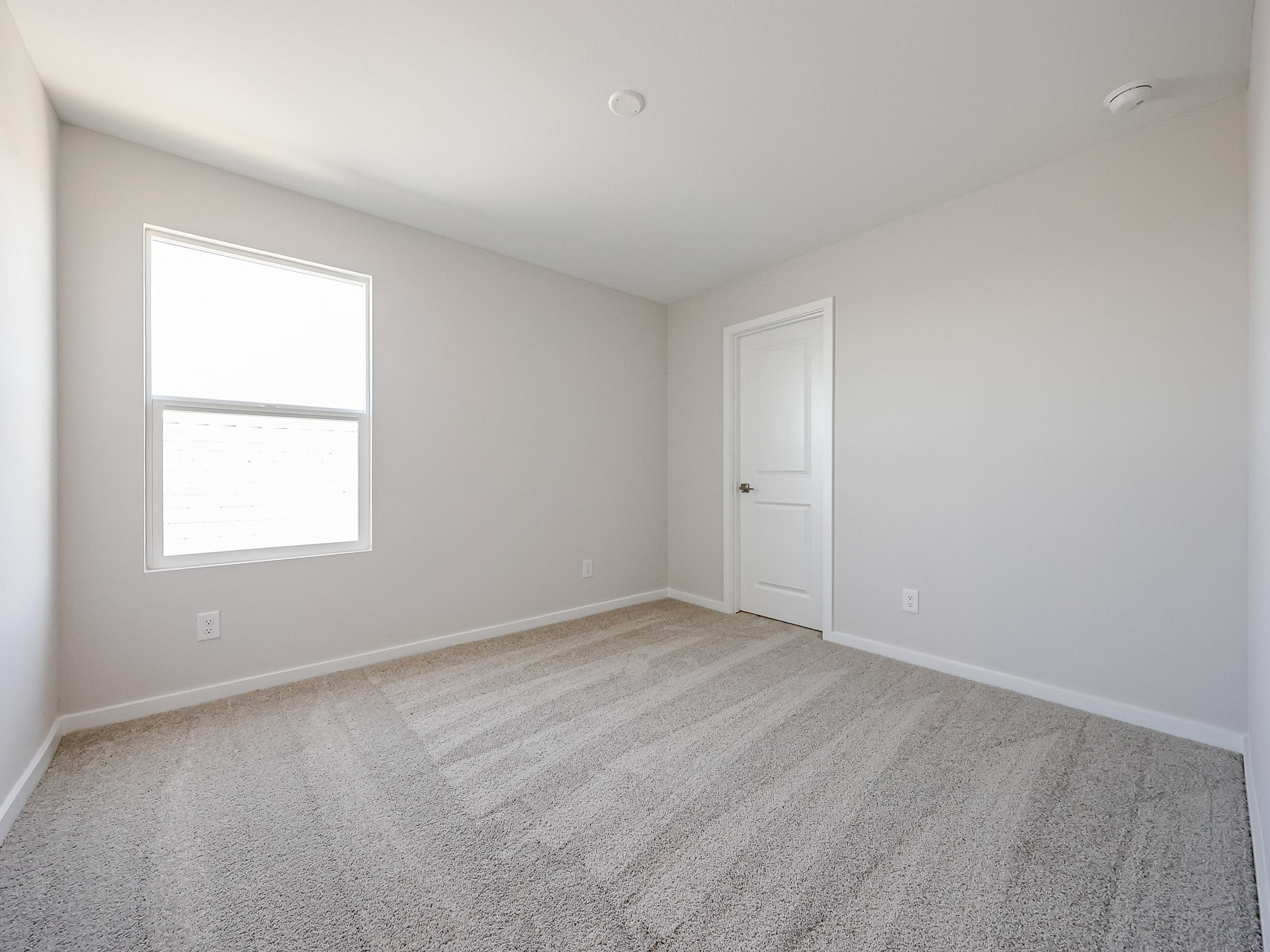 Empty bedroom with light-colored carpet, white walls, a closed white door, and a single window letting in natural light.