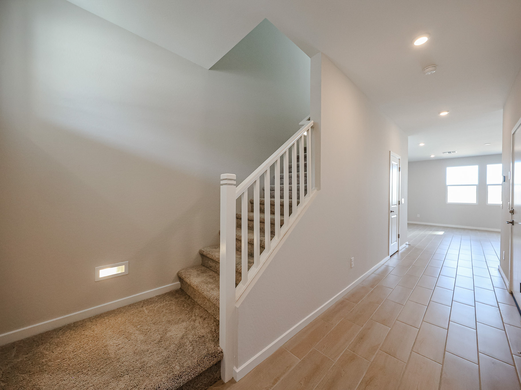 A modern home interior showing a carpeted staircase with a white railing on the left and a hallway with tile flooring leading to a bright room with windows.