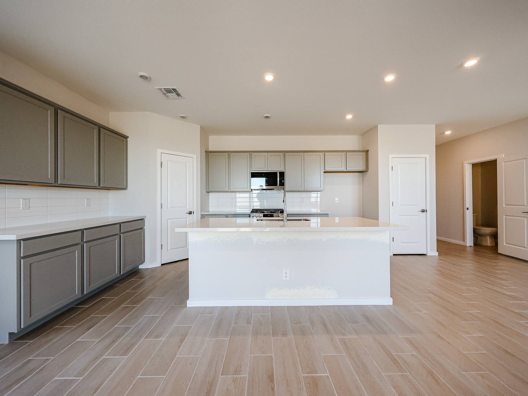 Modern kitchen with gray cabinets, stainless steel appliances, white countertops, light wood-look tile flooring, and recessed lighting. Two doors and an open pantry are visible.