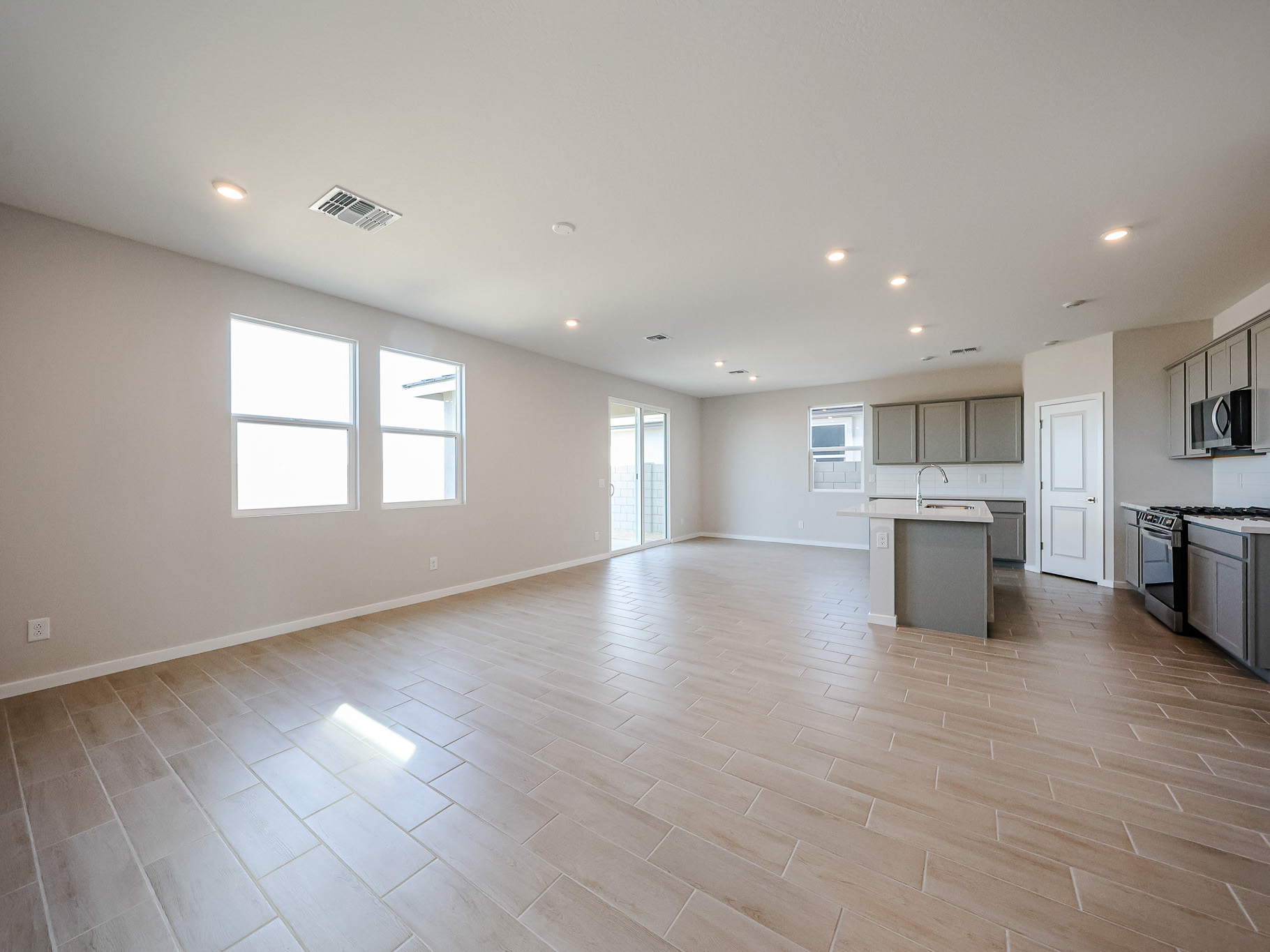 Spacious, empty open-plan living area with light wood-look tile flooring, recessed lighting, large windows, and a modern kitchen with gray cabinets and stainless steel appliances.