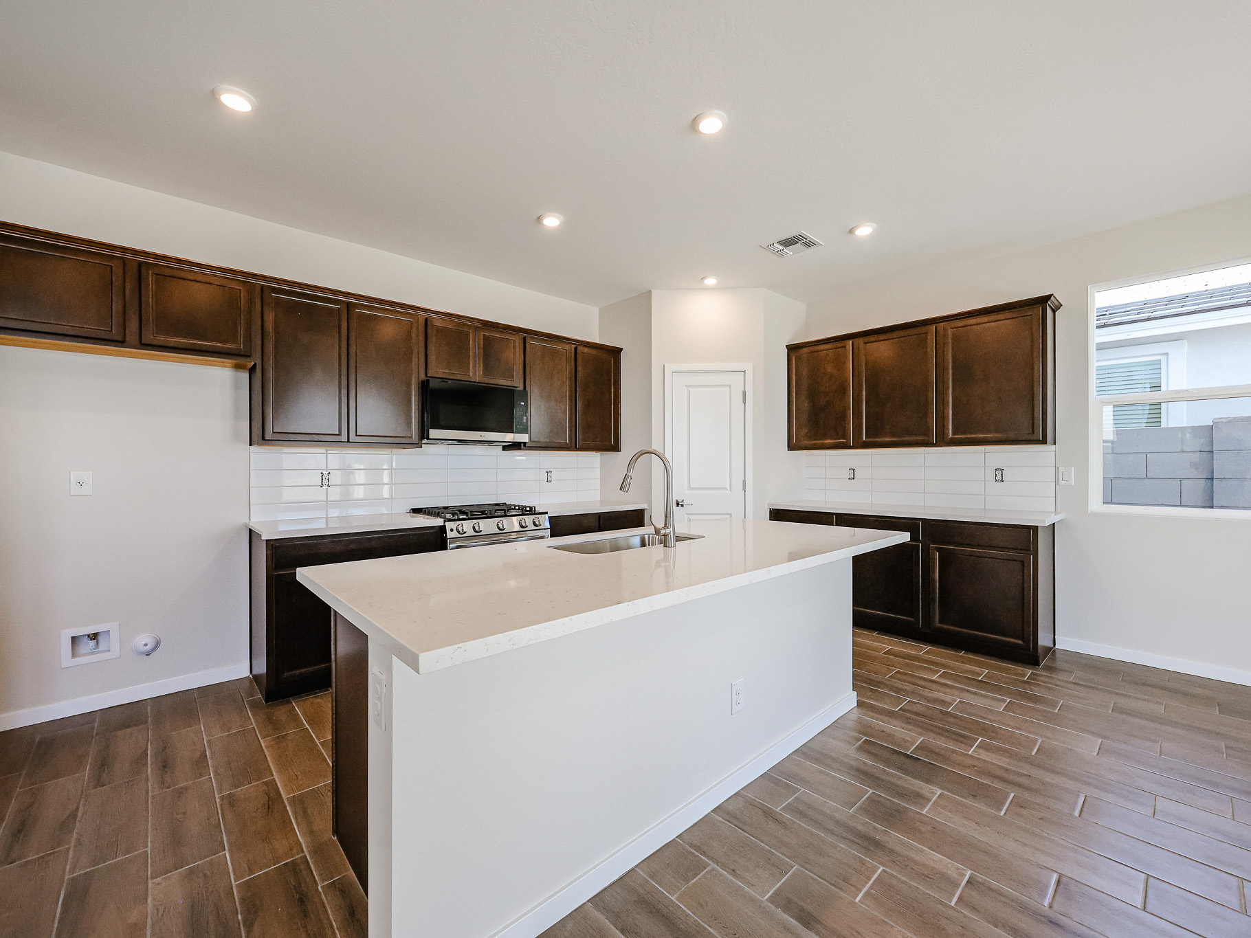 Modern kitchen with dark wood cabinets, white countertops, stainless steel appliances, tile flooring, and a central island. White backsplash and bright natural light from window.