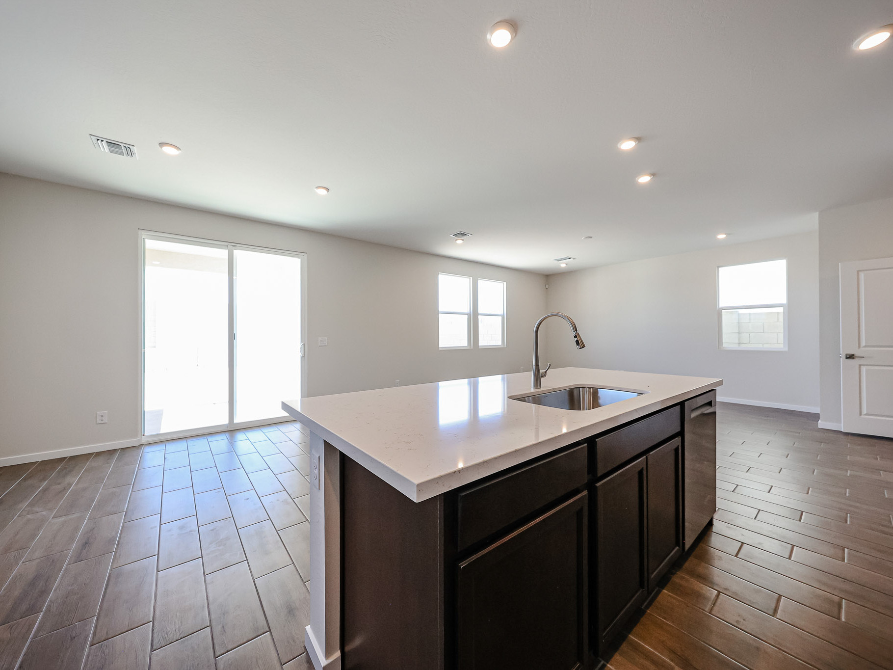 Modern empty kitchen and living area with tile flooring, white walls, recessed lighting, large windows, sliding glass door, and a central island with a sink.
