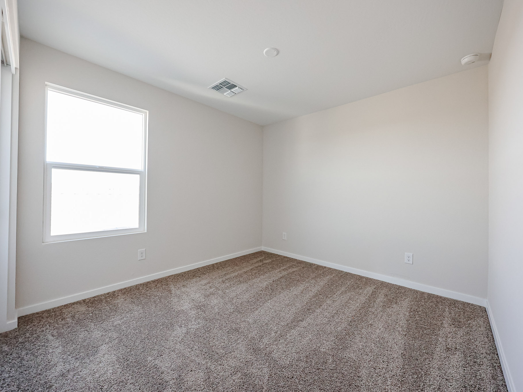 Empty room with beige carpet, off-white walls, a single window, and a ceiling vent. No furniture or decorations are present.