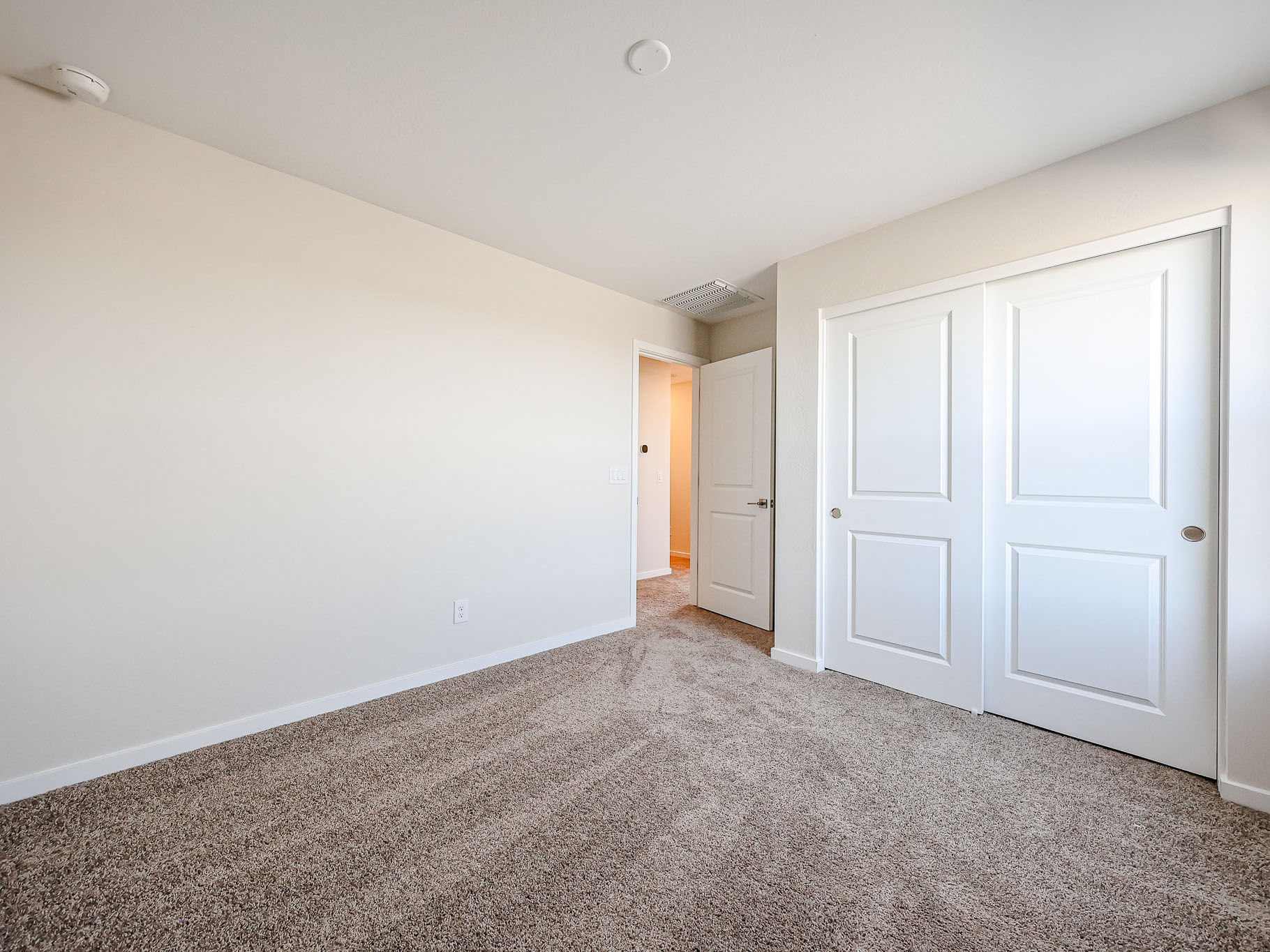 Empty bedroom with beige walls, carpeted floor, a closed double-door closet, and an open doorway leading to a lit hallway. Natural light enters from a window out of frame.